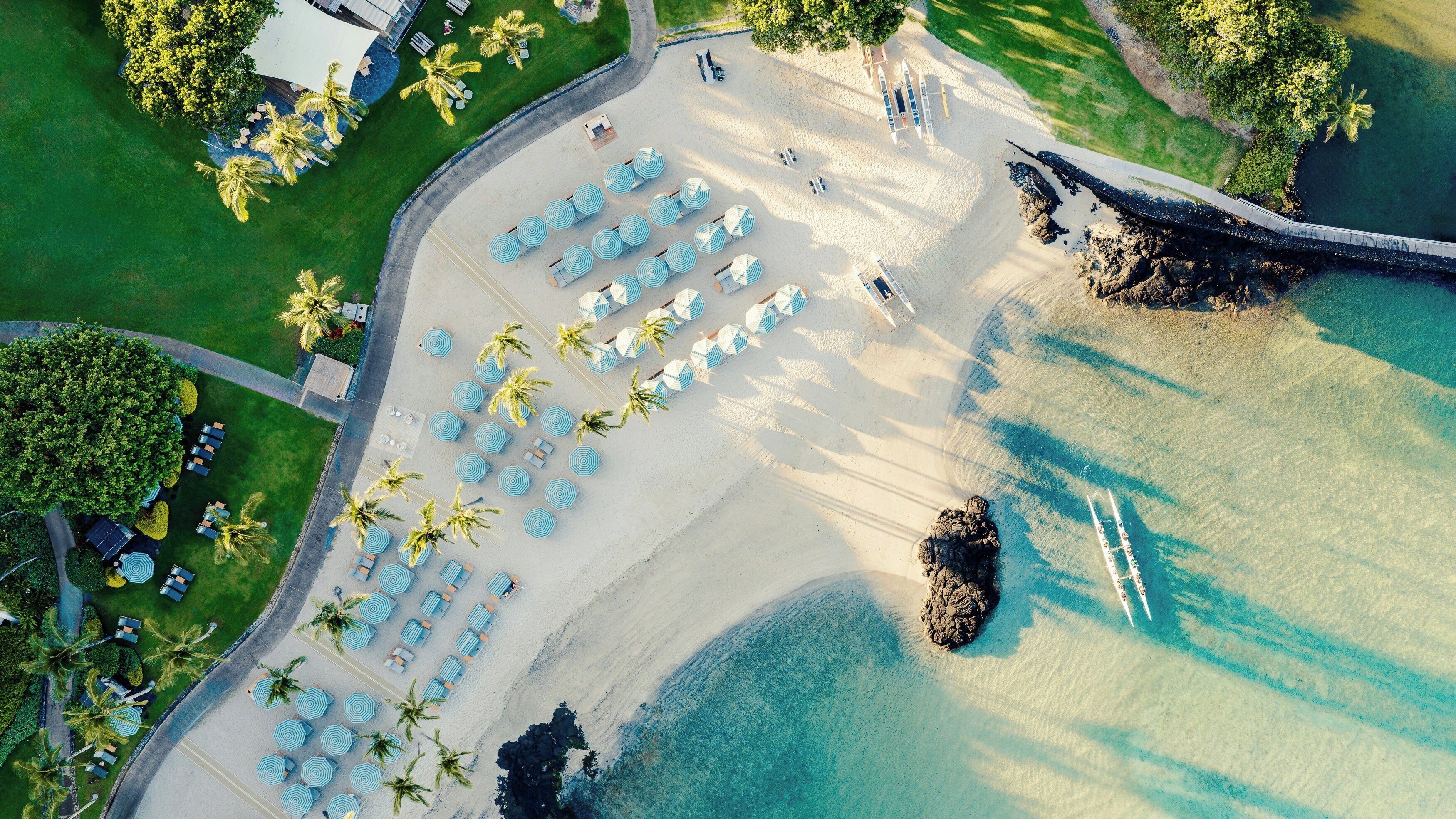 Vista aérea de un resort en la playa con tumbonas y sombrillas, un puente y un mar azul cristalino.