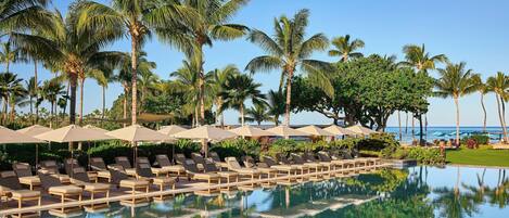A poolside area with lounge chairs and umbrellas, palm trees, and a clear view of the ocean.