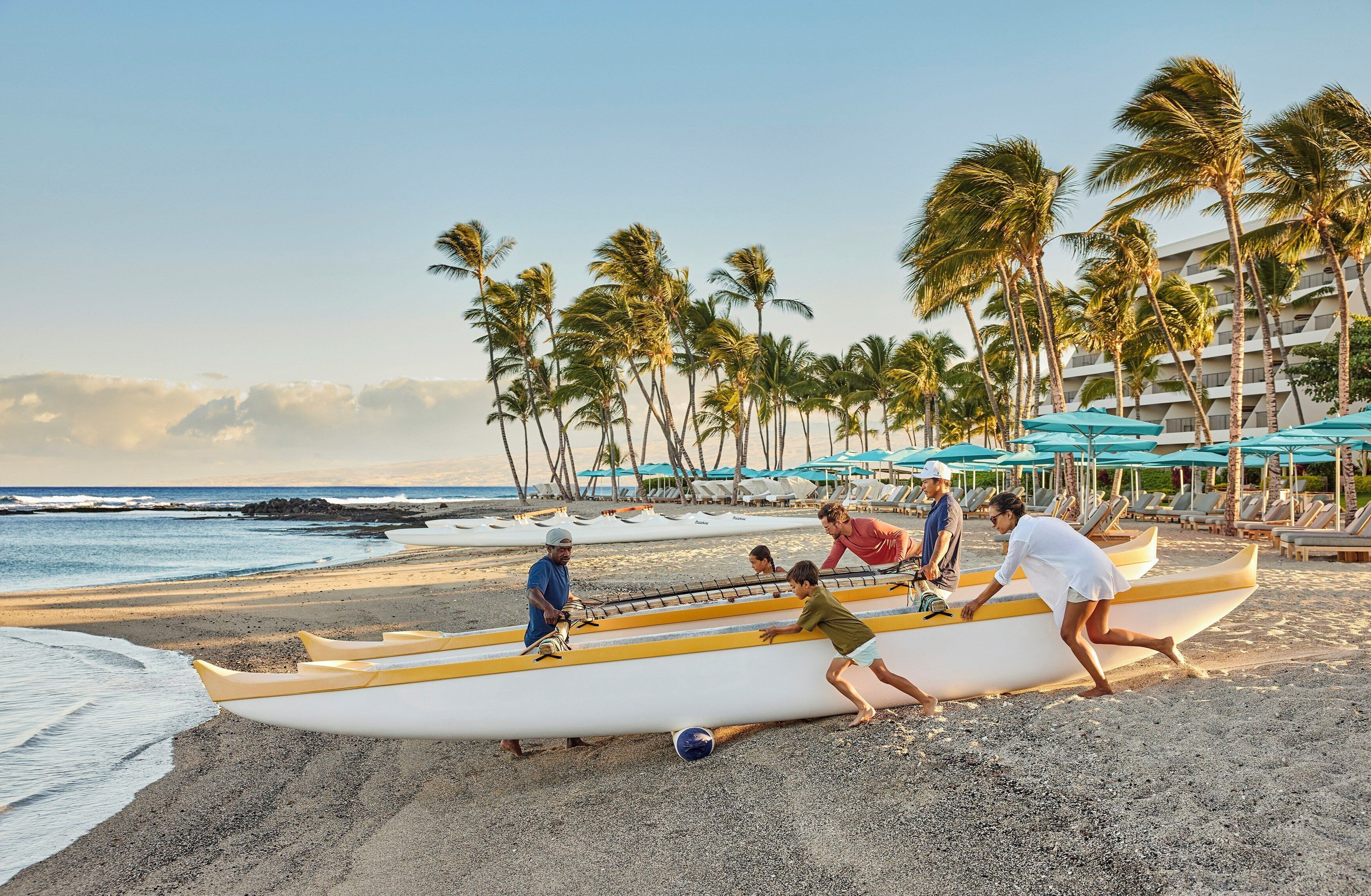 A beach with people handling a canoe, palm trees, and a hotel in the background.