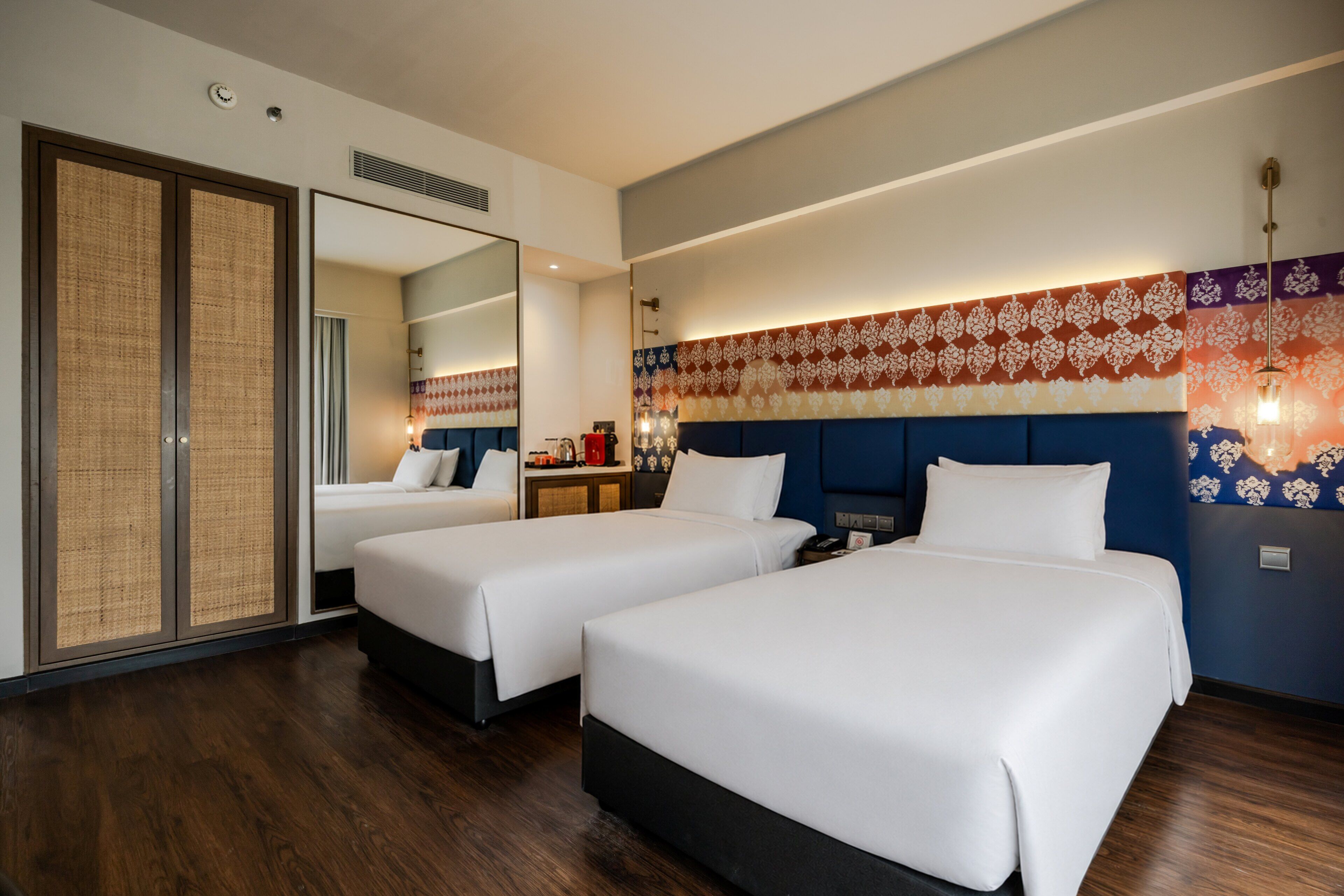 A hotel room with two beds, a large mirror, a wall-mounted light fixture, and a wooden floor.