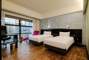 Une chambre d’hôtel avec deux lits, un bureau, une chaise et une vue sur la ville grâce à de grandes fenêtres.