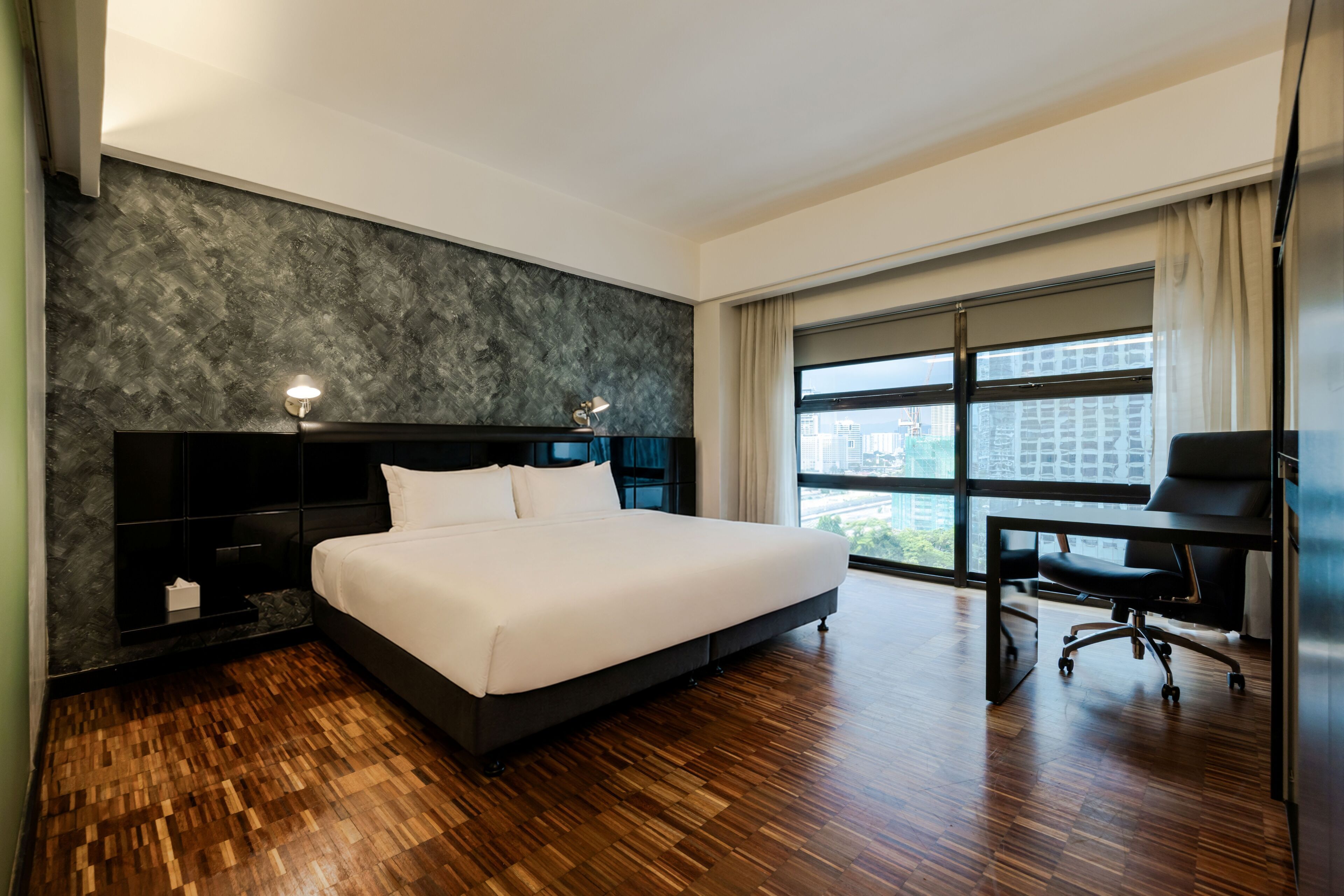 Una habitación de hotel moderna con una cama grande, un escritorio y vistas de la ciudad a través de amplios ventanales.