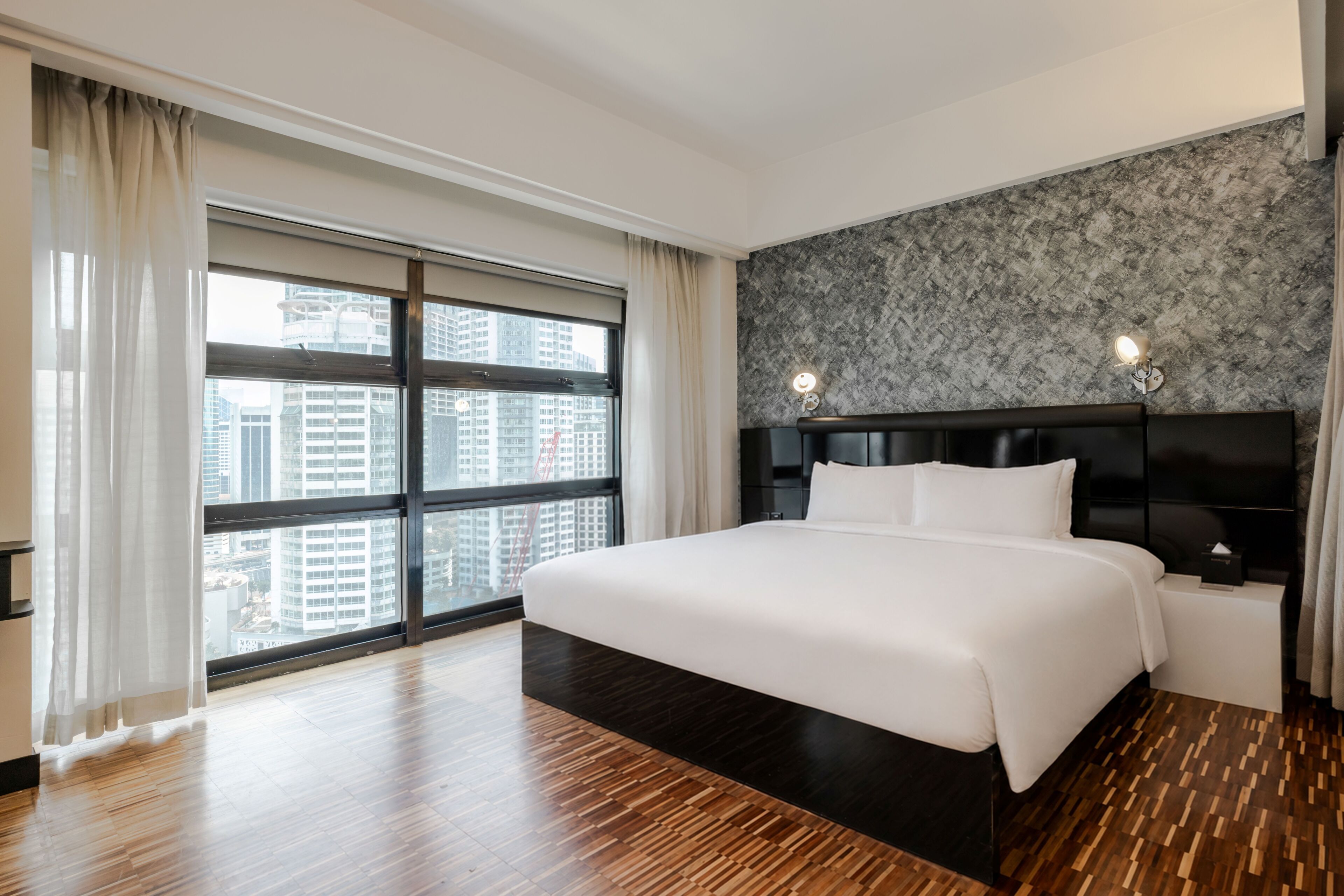 Habitación de hotel con una cama grande, vista de la ciudad a través de ventanales y una pared con textura.