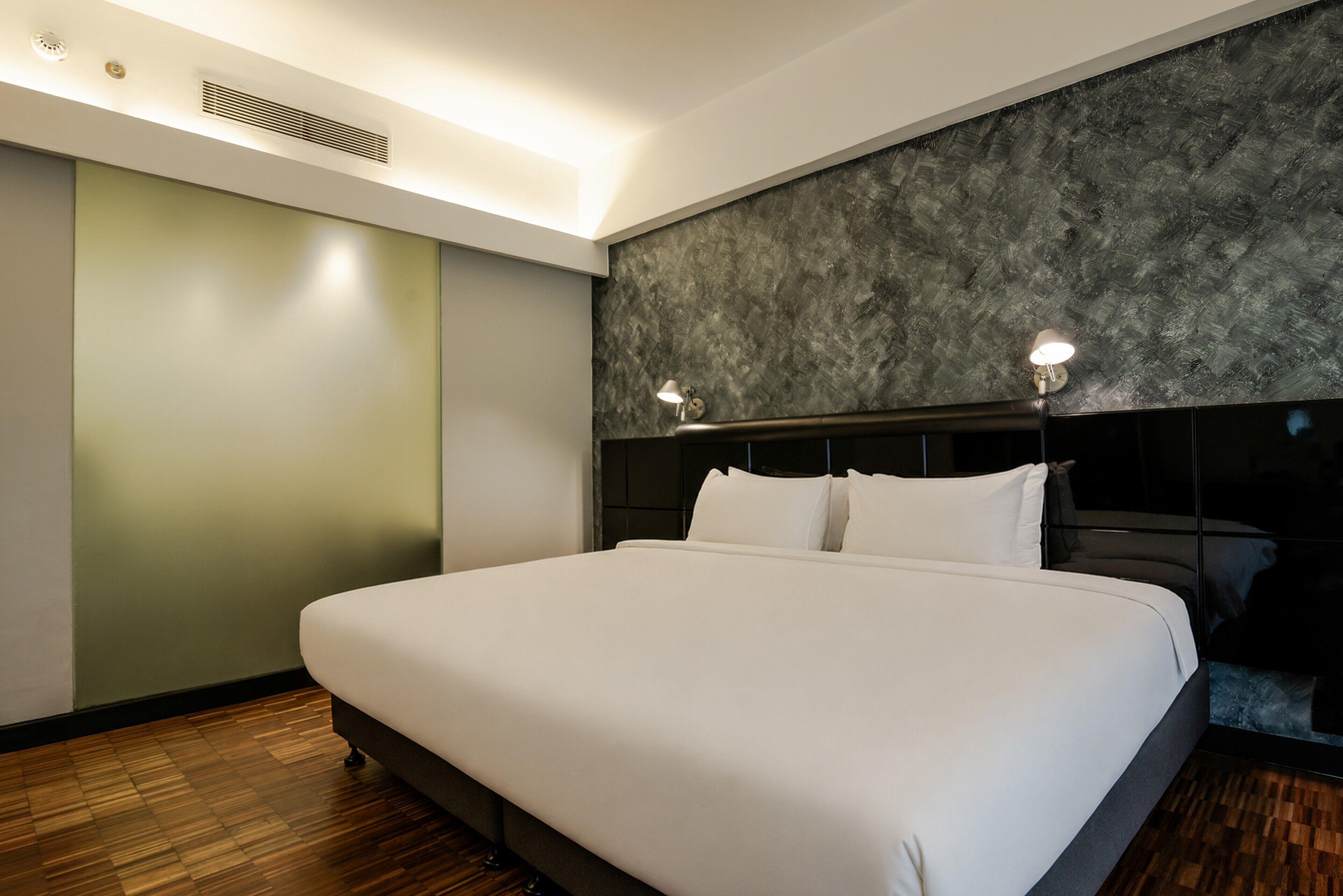 A modern hotel room with a large bed, a headboard with integrated lighting, a wooden floor, and a textured wall.