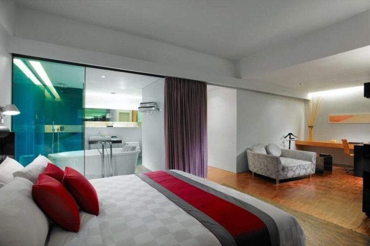 A modern hotel room with a large bed, a desk, a chair, and a view of the kitchen.