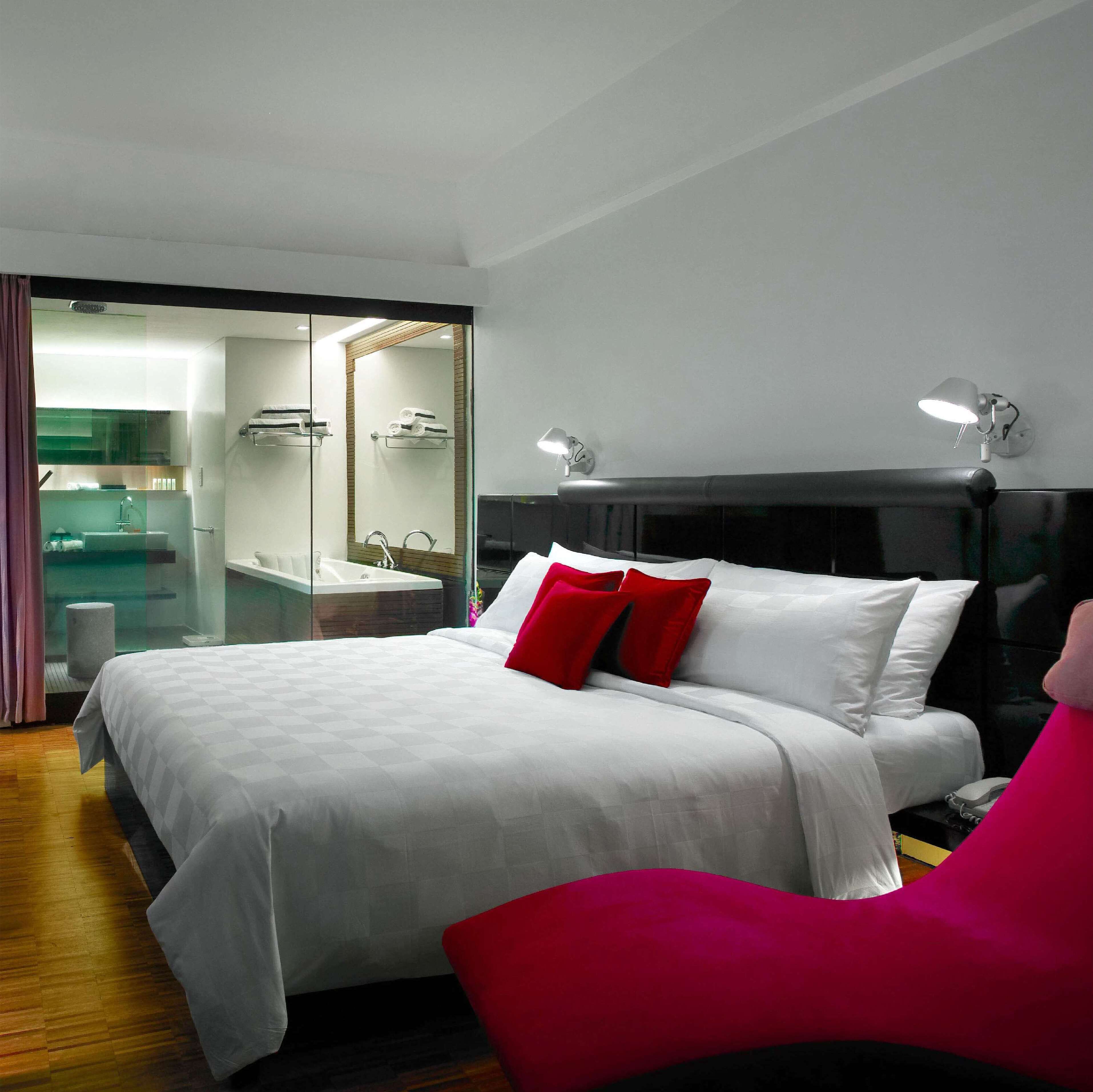 Une chambre d’hôtel moderne avec un grand lit, une salle de bain lumineuse dotée d’une douche en verre et un fauteuil rouge.