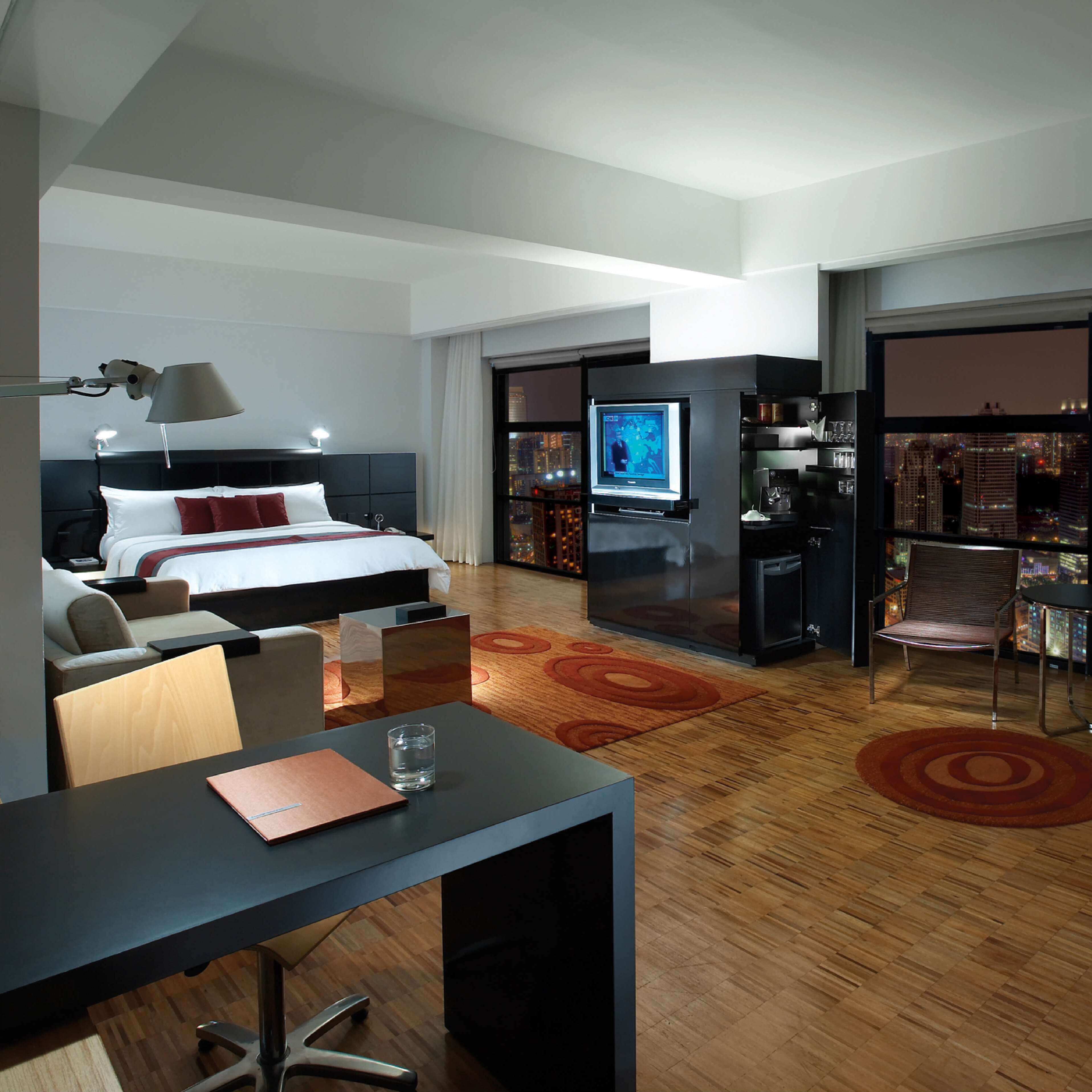 A modern hotel room with a bed, desk, chair, TV, and a minibar.