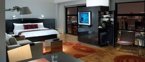 A modern hotel room with a bed, desk, chair, TV, and a minibar.