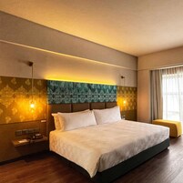 A modern hotel room with a large bed, a headboard with a textured design, a bedside table with a lamp, and a window with curtains.