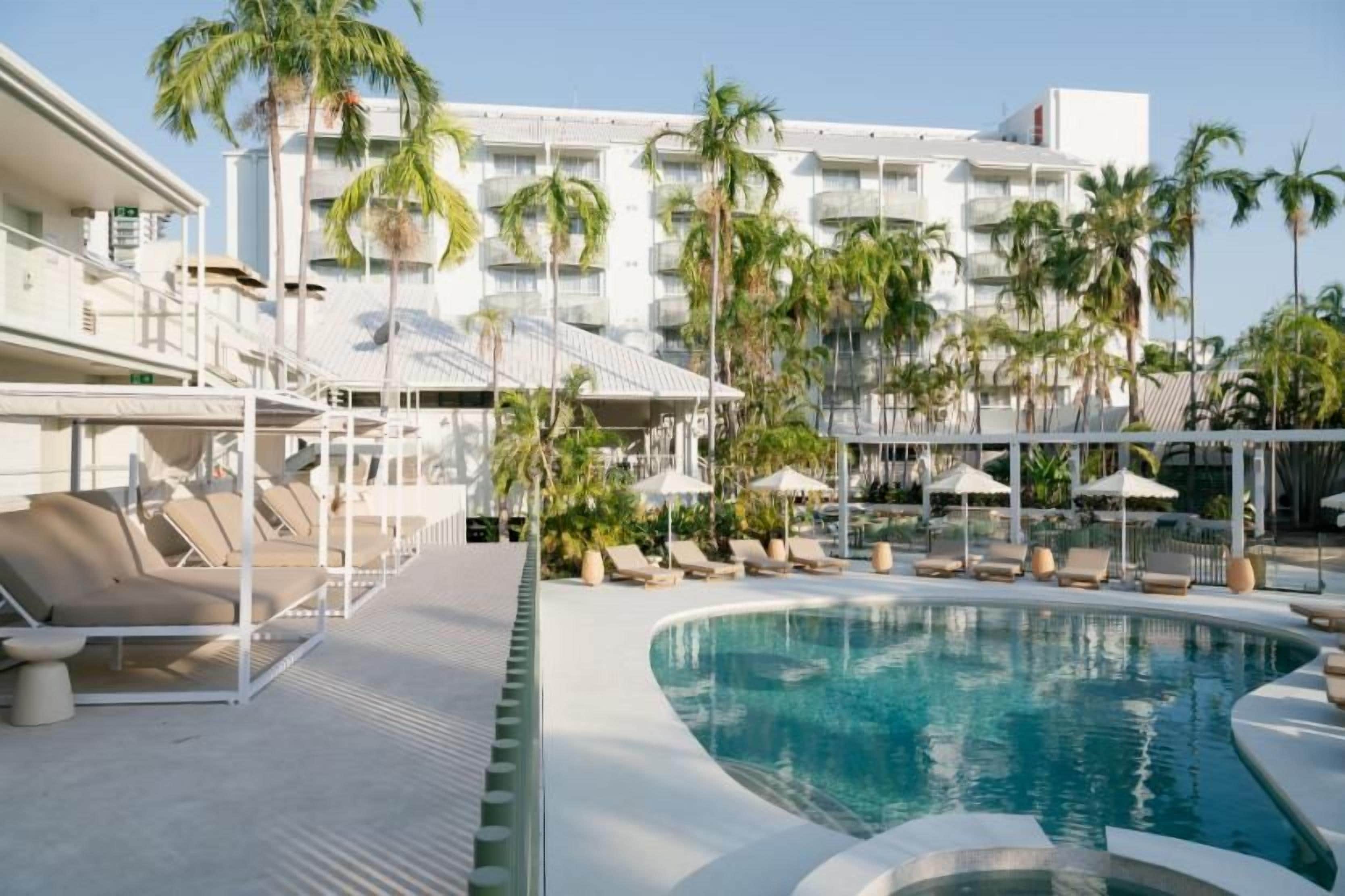 A pool area with lounge chairs and umbrellas, surrounded by palm trees and a hotel building in the background.