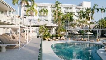 A pool area with lounge chairs and umbrellas, surrounded by palm trees and a hotel building in the background.