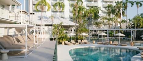 A pool area with lounge chairs and umbrellas, surrounded by palm trees and a hotel building in the background.