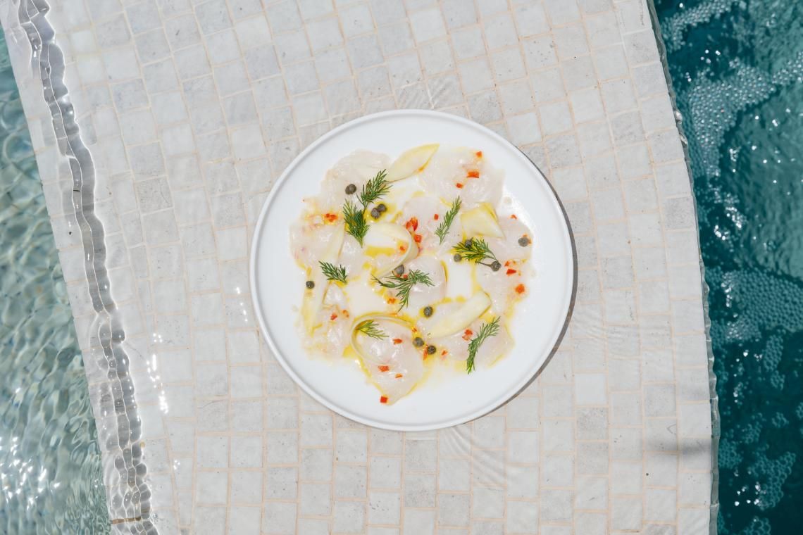 A white plate with a seafood dish, garnished with dill and capers, placed on a tiled surface near a pool.