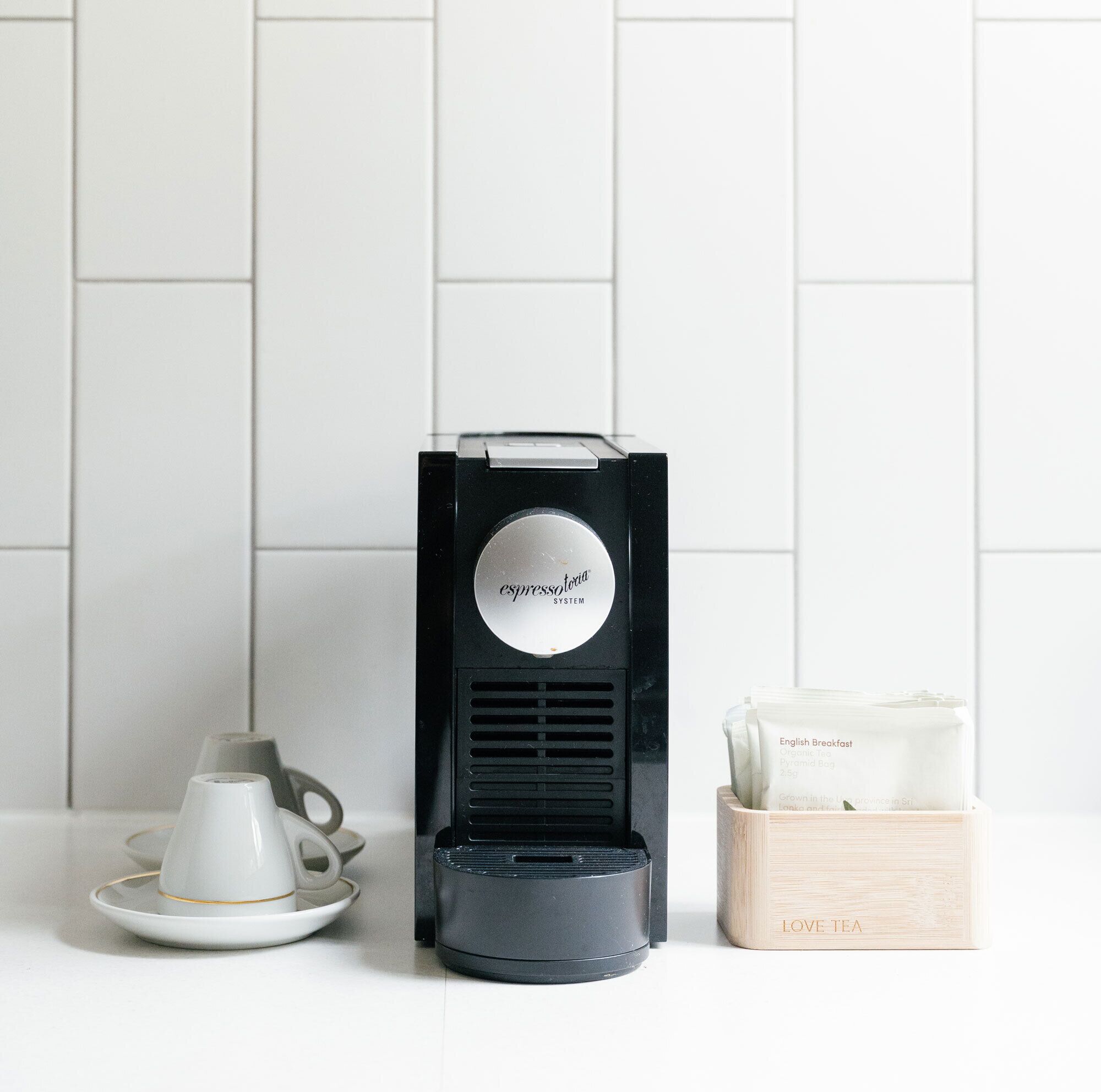 Une machine à café, une théière et une boîte de thé sont posés sur un plan de travail blanc.