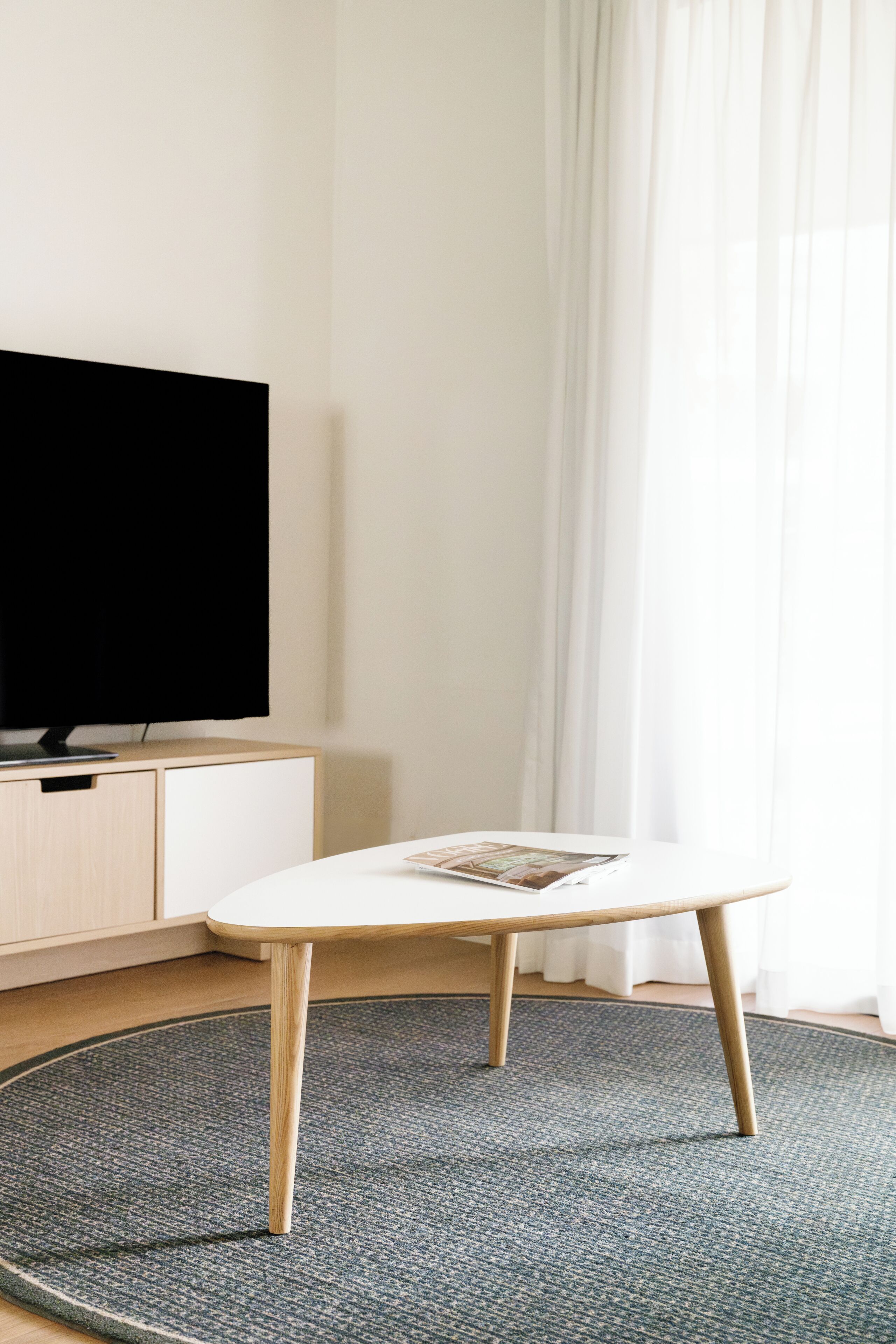 Quarto com televisão sobre um armário de madeira, uma mesa redonda com uma revista e uma janela com cortinas leves.