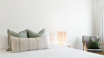 A neatly made bed with pillows, a bedside lamp, and a chair in a room with white walls and a wooden headboard.