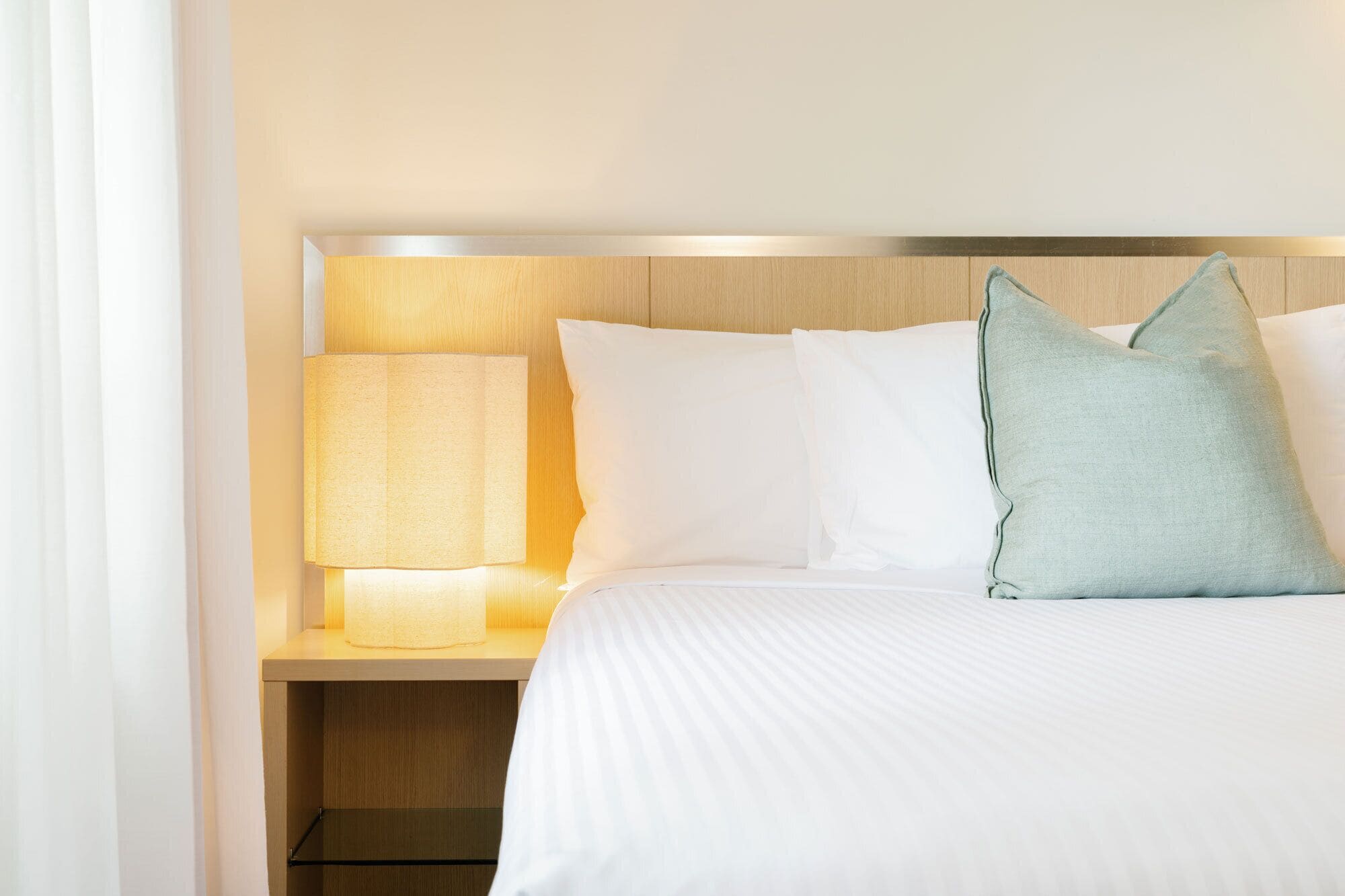 Een hotelkamer met een bed, witte lakens, een nachtkastje met een lamp en een houten hoofdeinde.