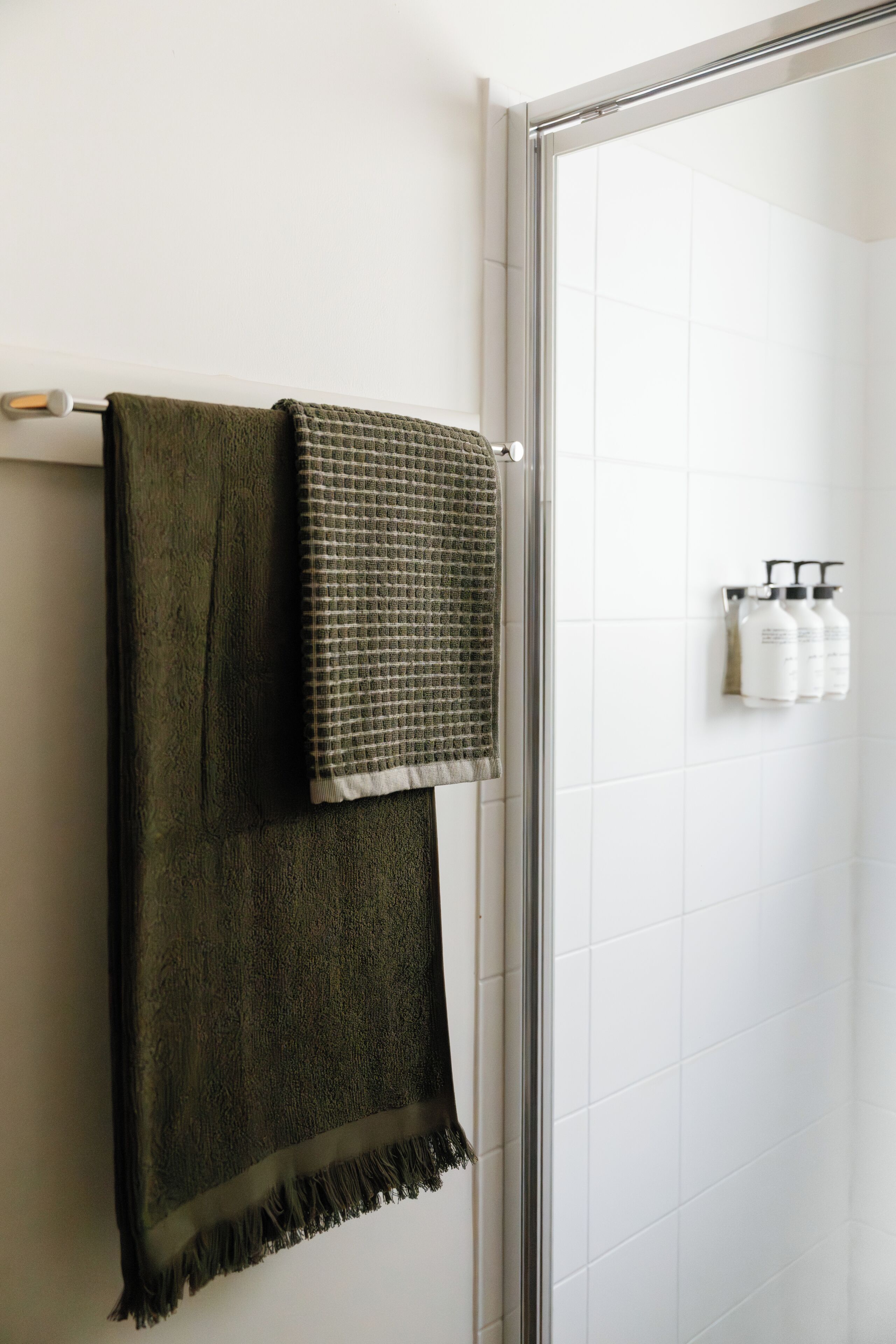 Un baño con un perchero que sostiene dos toallas, una puerta de ducha de vidrio y un dispensador de botellas en la pared.