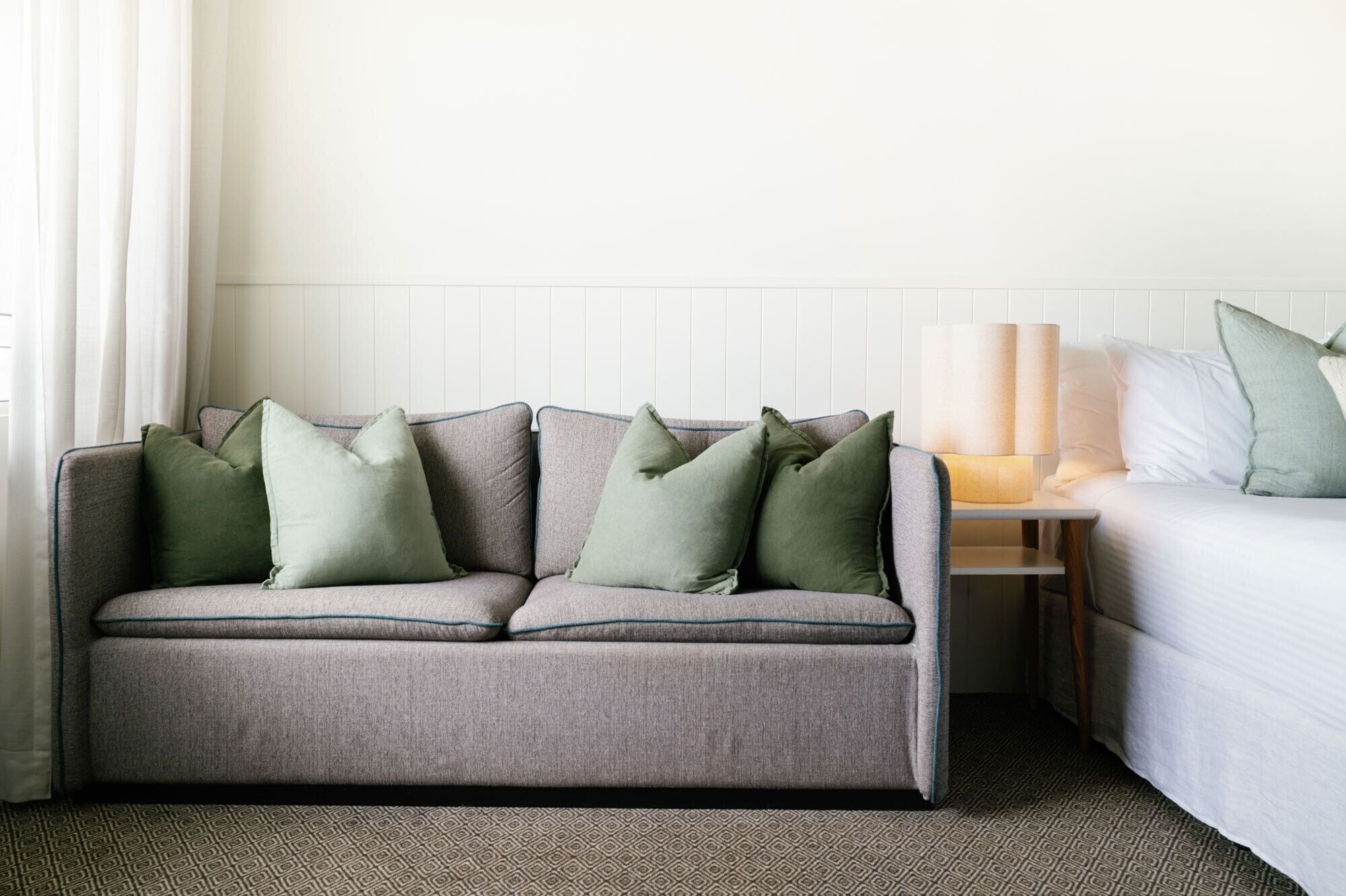 A neutral-colored sofa with green pillows, a bedside table with a lamp, and a white bed in a hotel room.