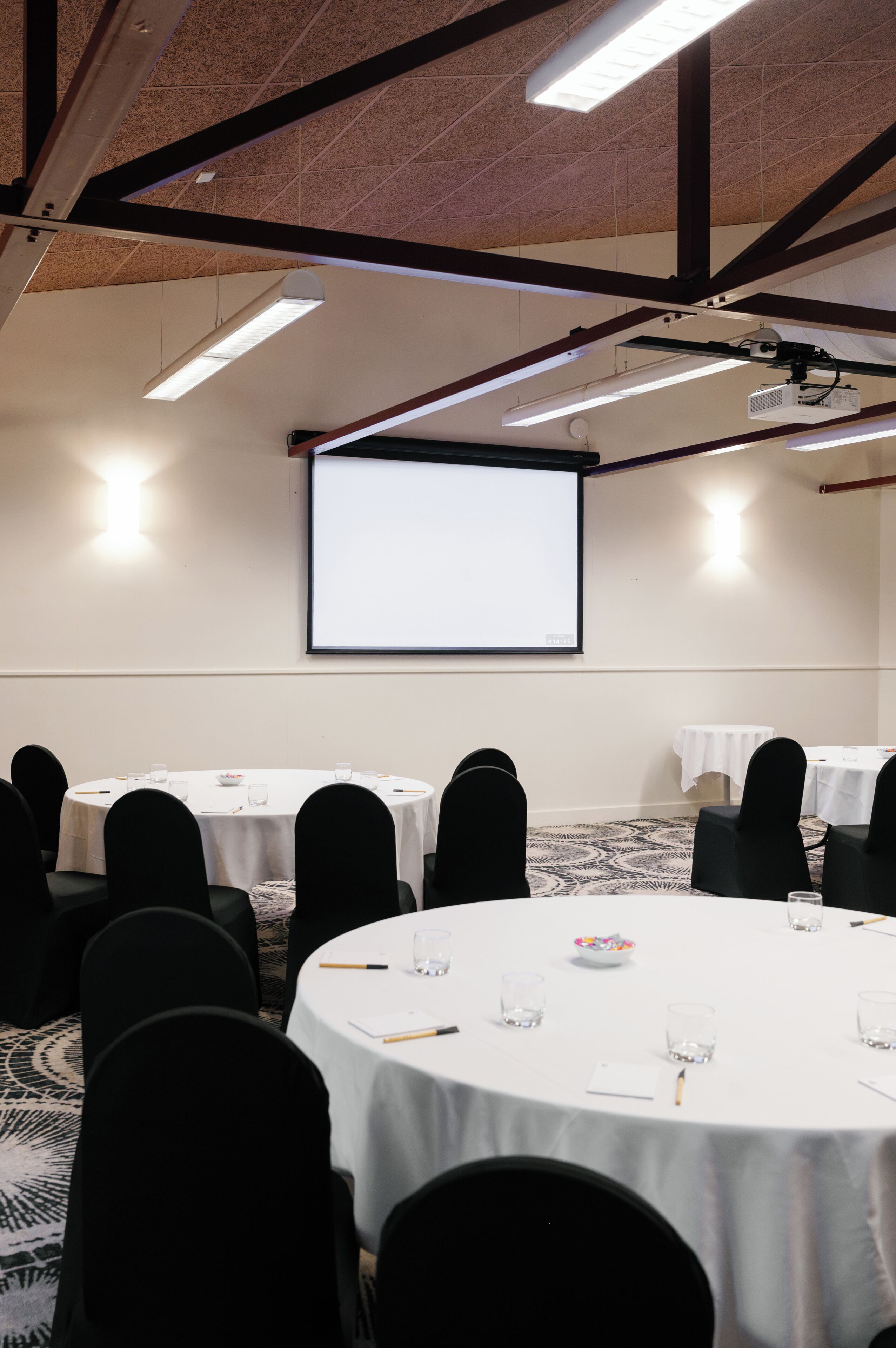 A conference room with round tables, black chairs, a projector screen, and a carpeted floor.