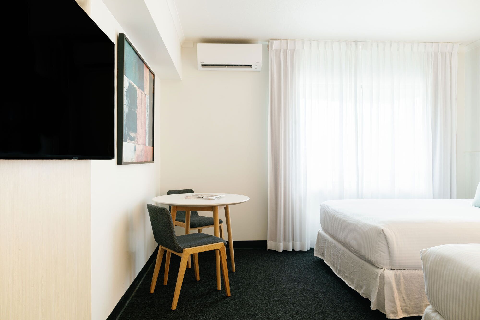Habitación de hotel con cama, una pequeña mesa con dos sillas, un televisor de pantalla plana montado en la pared y un equipo de aire acondicionado.