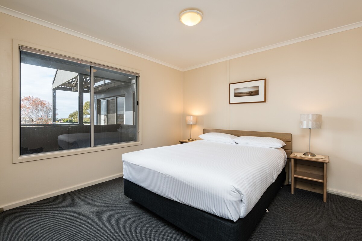 A hotel room with a large window, a bed, a nightstand with a lamp, and a framed picture on the wall.