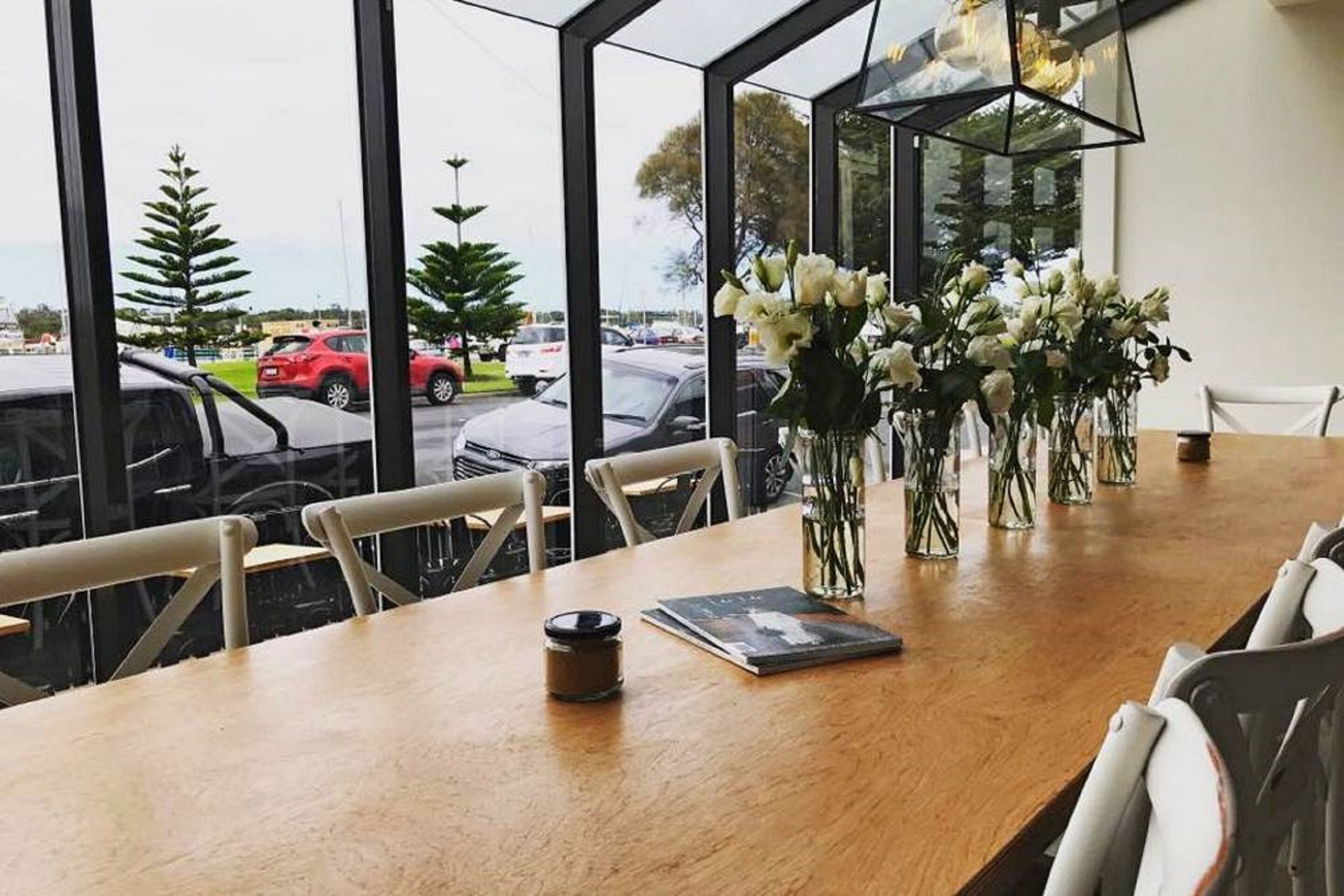 A dining area with a wooden table, white chairs, and a view of parked cars through large windows.