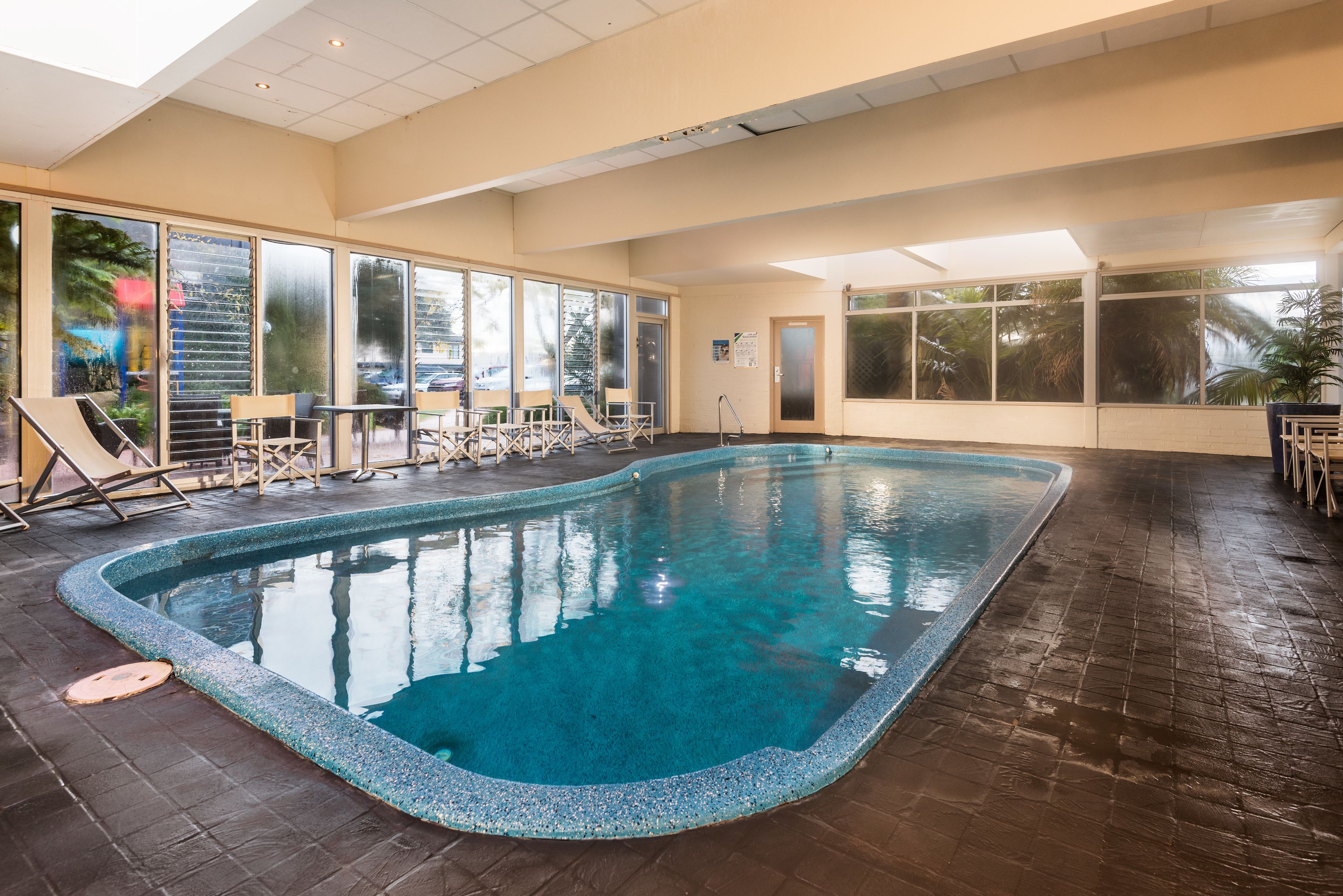 An indoor swimming pool with clear blue water, surrounded by lounge chairs and tables, large windows with views of palm trees, and a tiled floor.