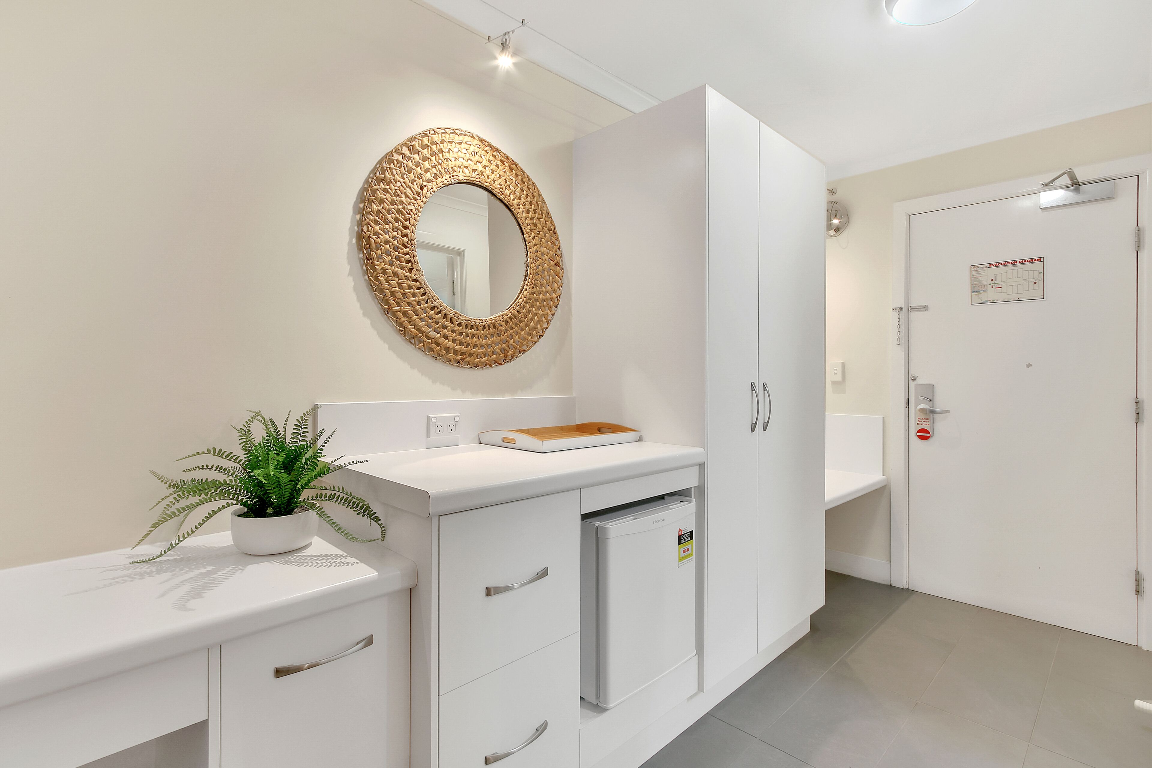 Un baño moderno con un tocador blanco, un espejo redondo, un refrigerador pequeño y una planta en maceta.
