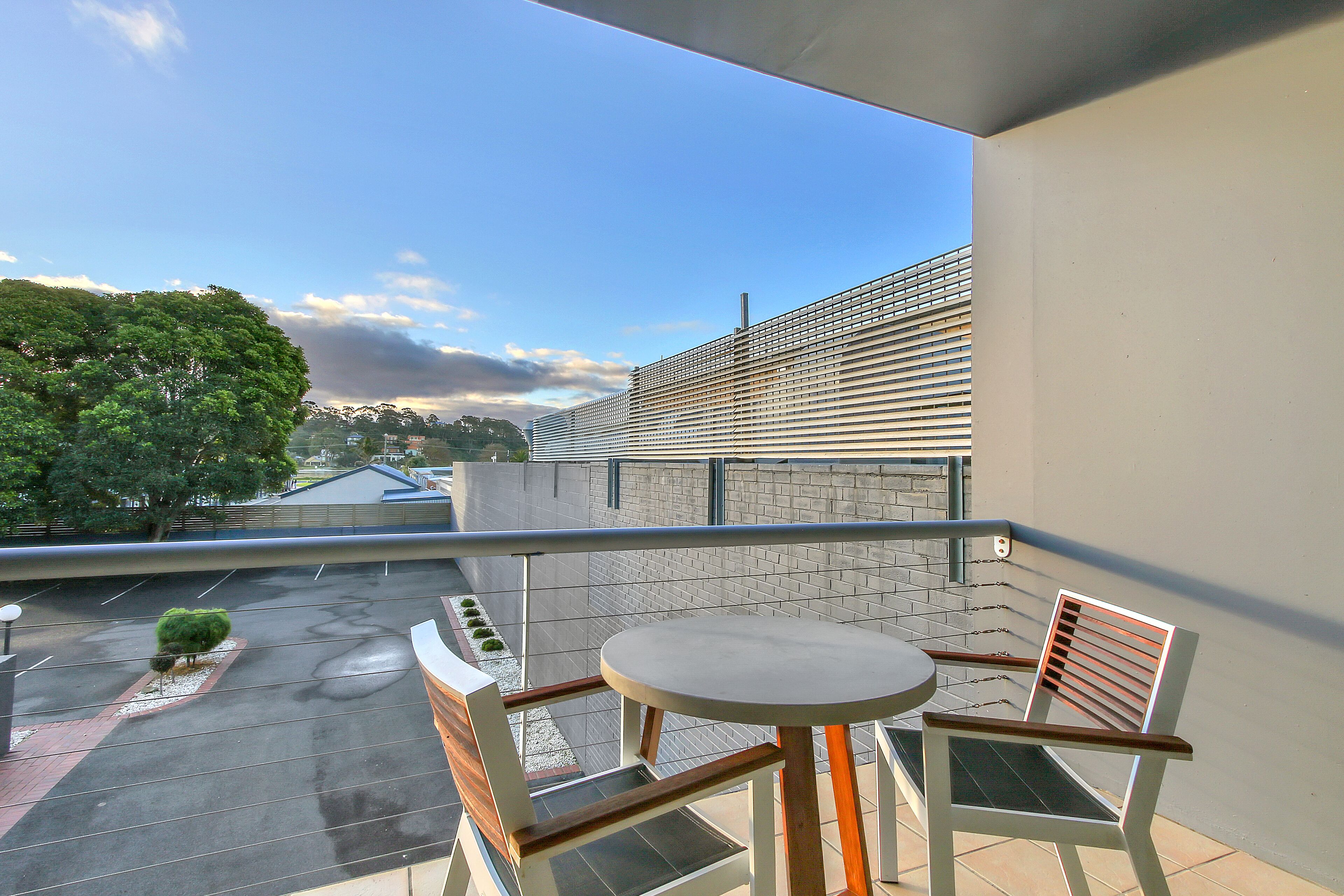 Un balcón con mesa y sillas, vista a un edificio de ladrillo y un árbol al fondo.