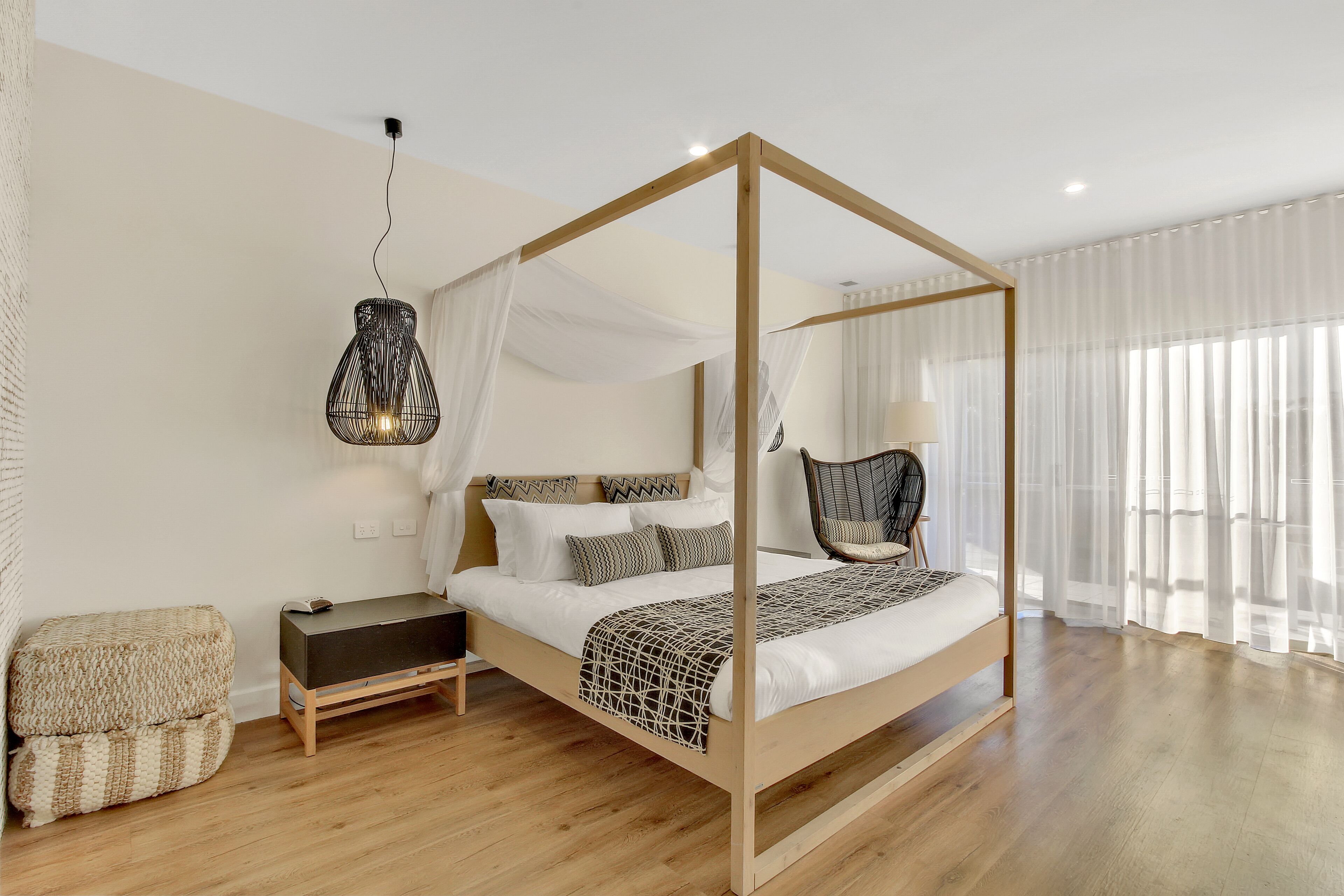 Een kamer met een hemelbed, nachtkastje, stoel en hanglamp, met houten vloer en dunne gordijnen.