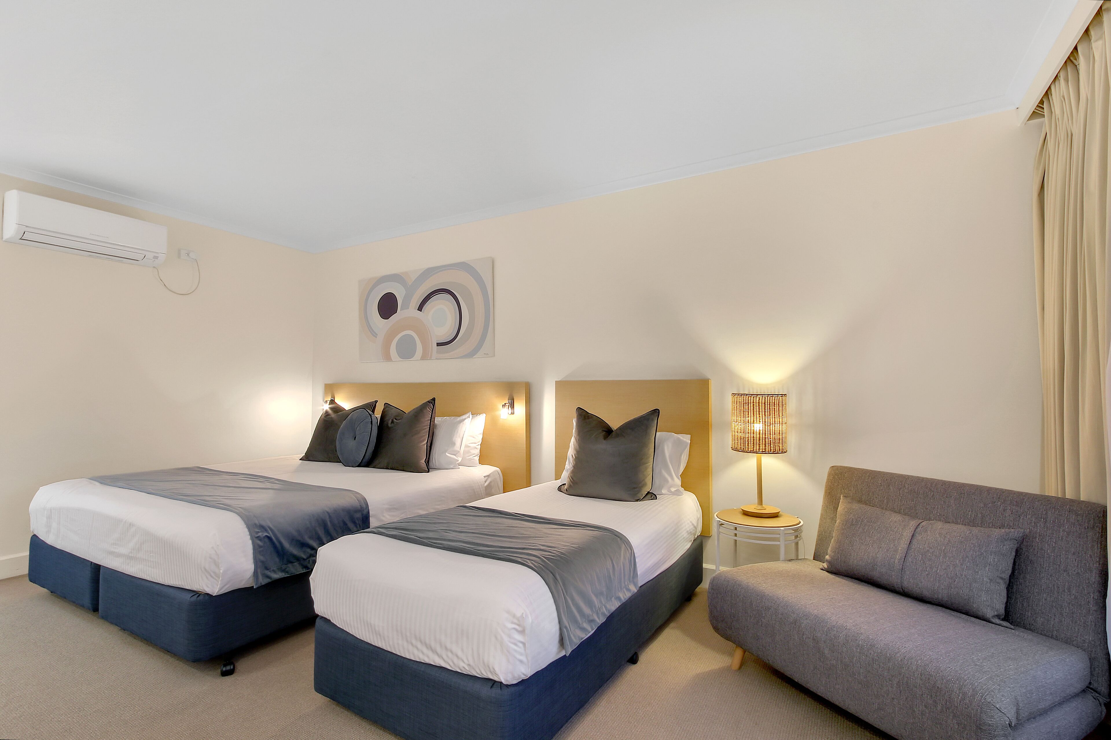 Une chambre d’hôtel avec deux lits, un canapé, une lampe et un climatiseur fixé au mur.