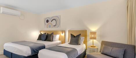 A hotel room with two beds, a sofa, a lamp, and a wall-mounted air conditioner.