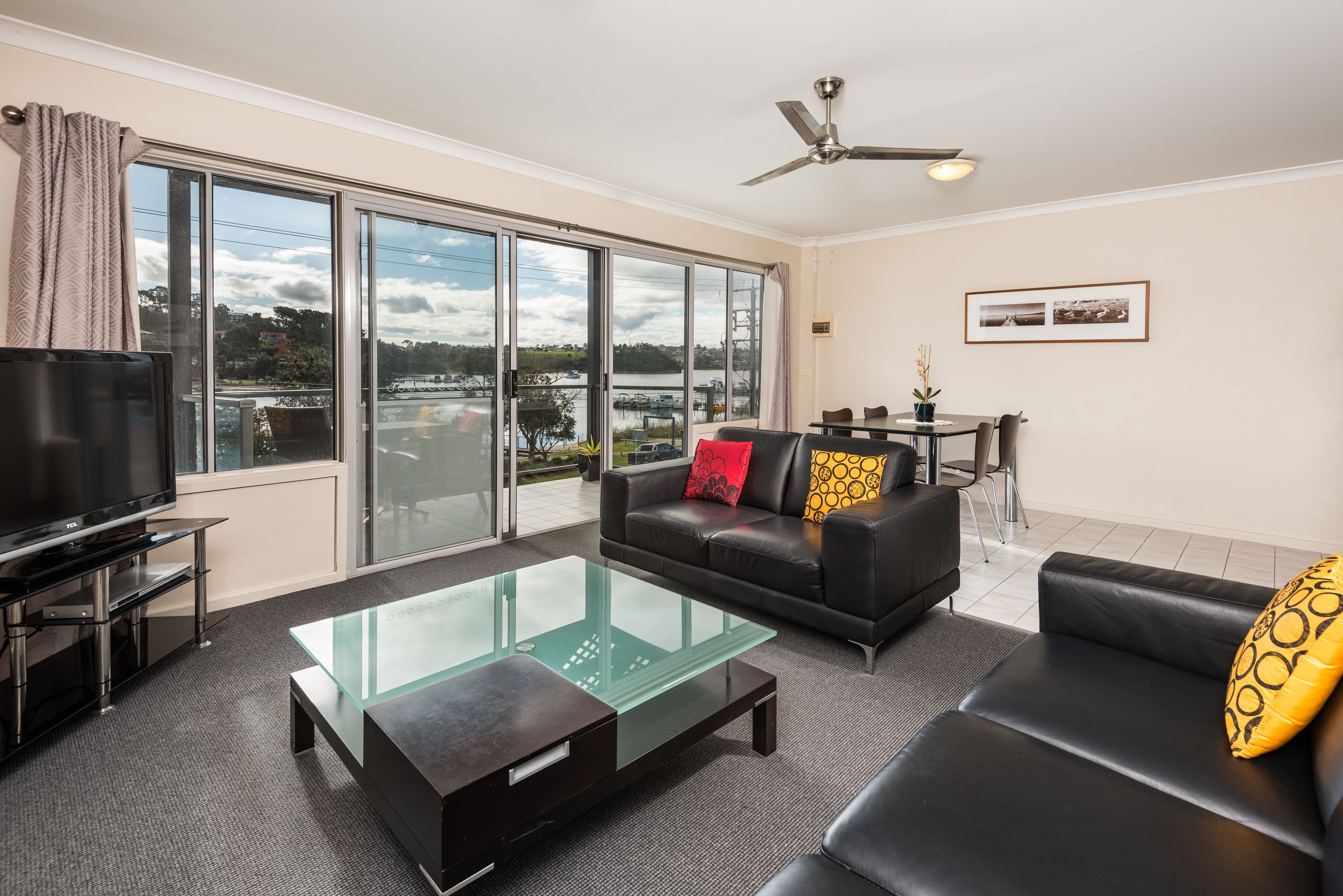 A modern living room with a glass coffee table, black sofas, a TV, and a view of a lake through large windows.
