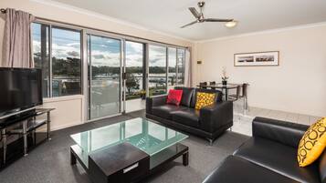 A modern living room with a glass coffee table, black sofas, a TV, and a view of a lake through large windows.