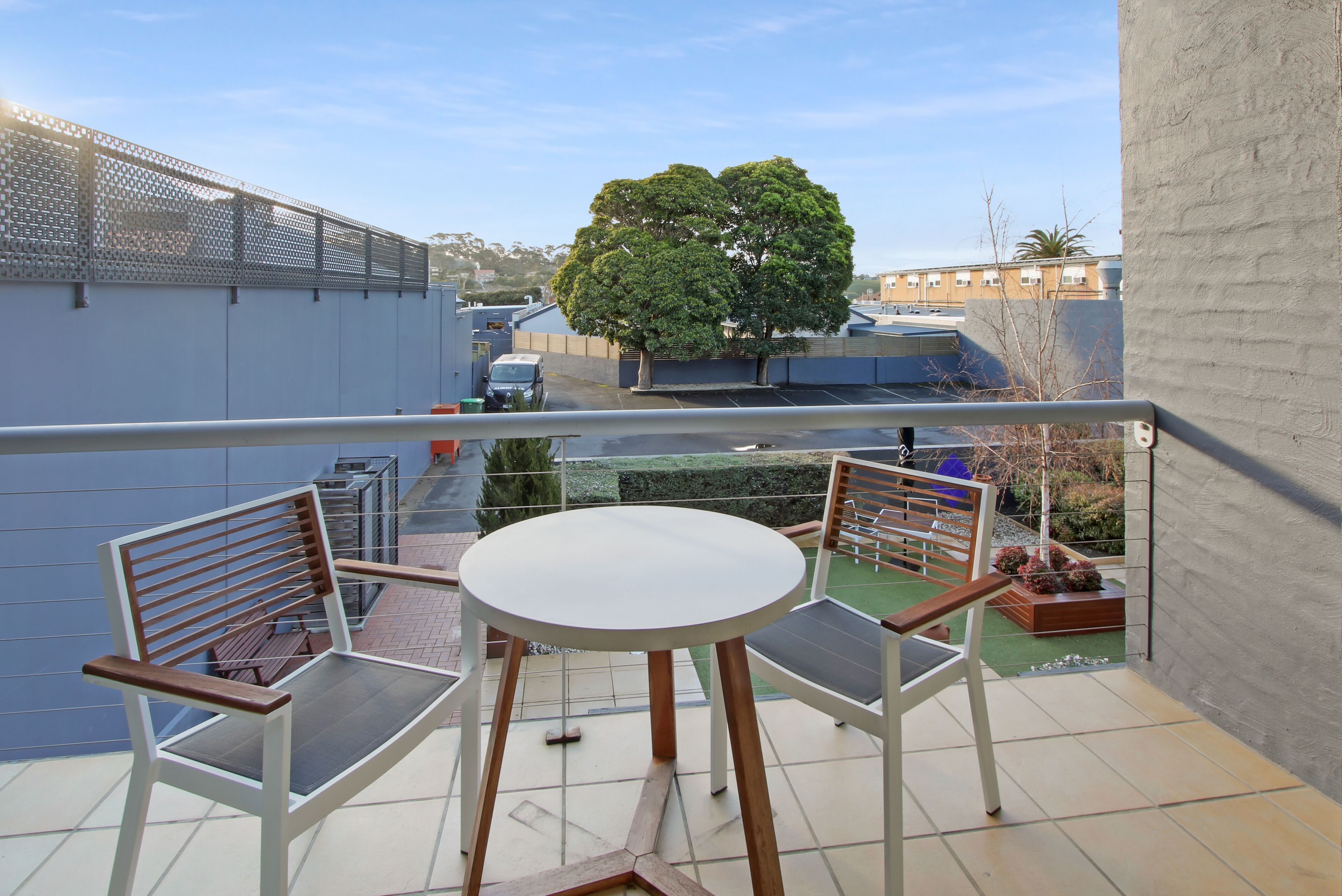 Ein Balkon mit rundem Tisch und zwei Stühlen, Blick auf einen Baum und ein Gebäude im Hintergrund.