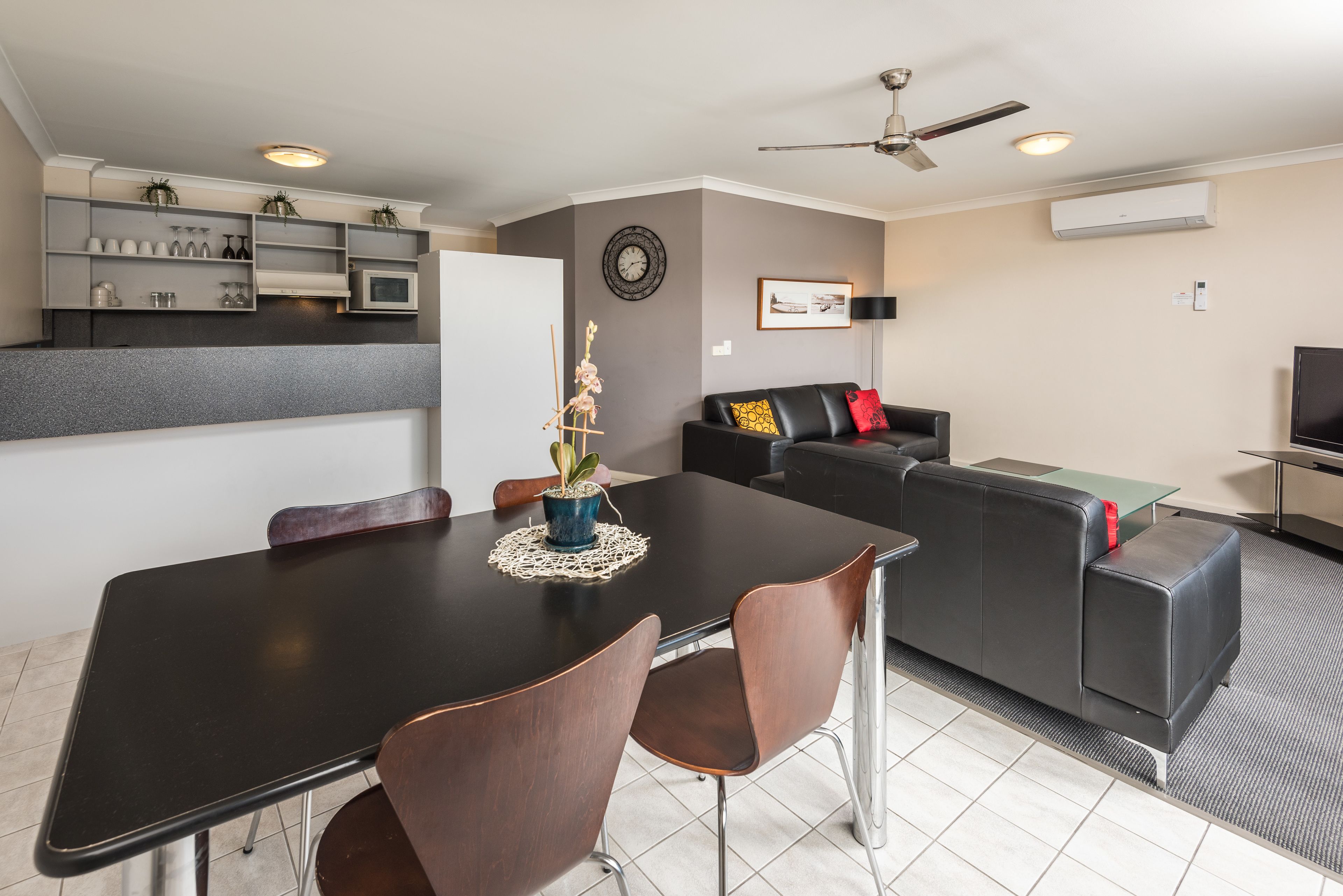 Una sala moderna con una mesa de comedor negra, sillas marrones, un sofá negro con cojines amarillos y rojos, un ventilador de techo y una zona de cocina con estantes y un microondas.