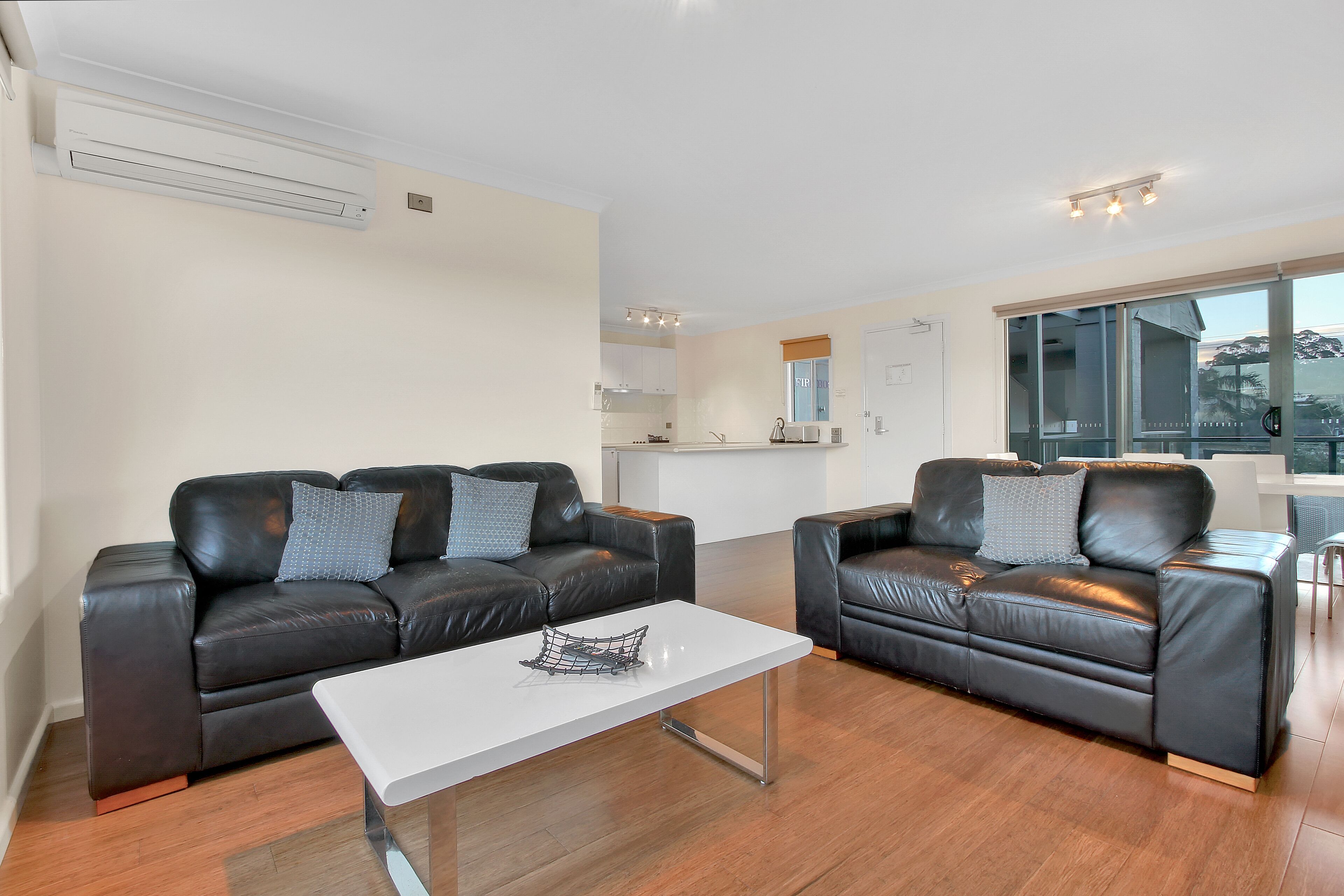 A modern living room with two black leather sofas, a white coffee table, and a kitchen in the background.