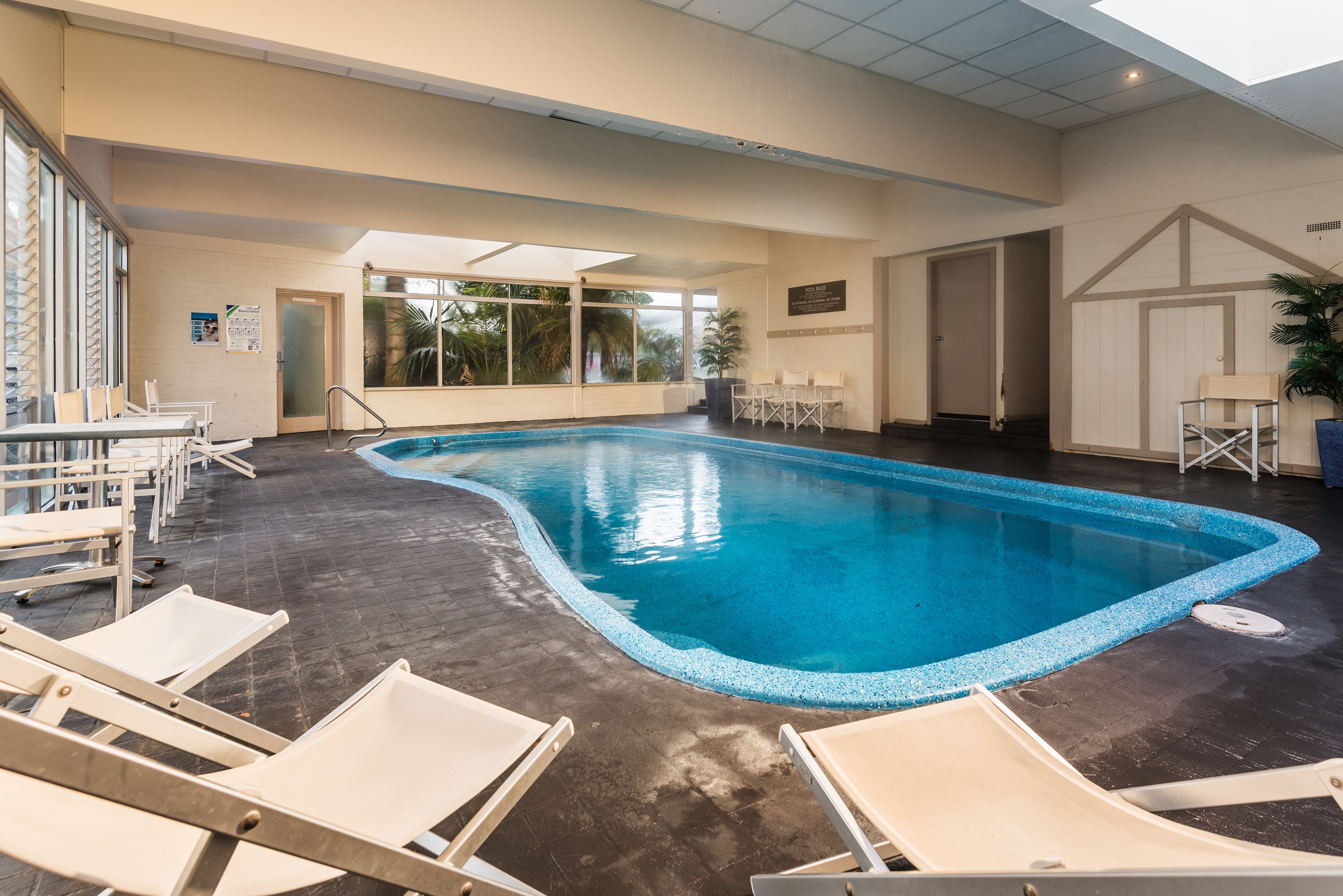 An indoor swimming pool with clear blue water, surrounded by white lounge chairs and a tiled floor.