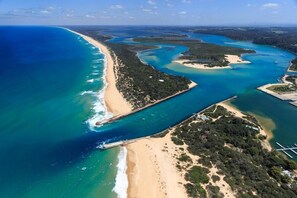 An aerial view of a coastal area with a beach, clear blue waters, and a mix of sandy and vegetated areas. - Bellevue on the Lakes (Lakes Entrance)