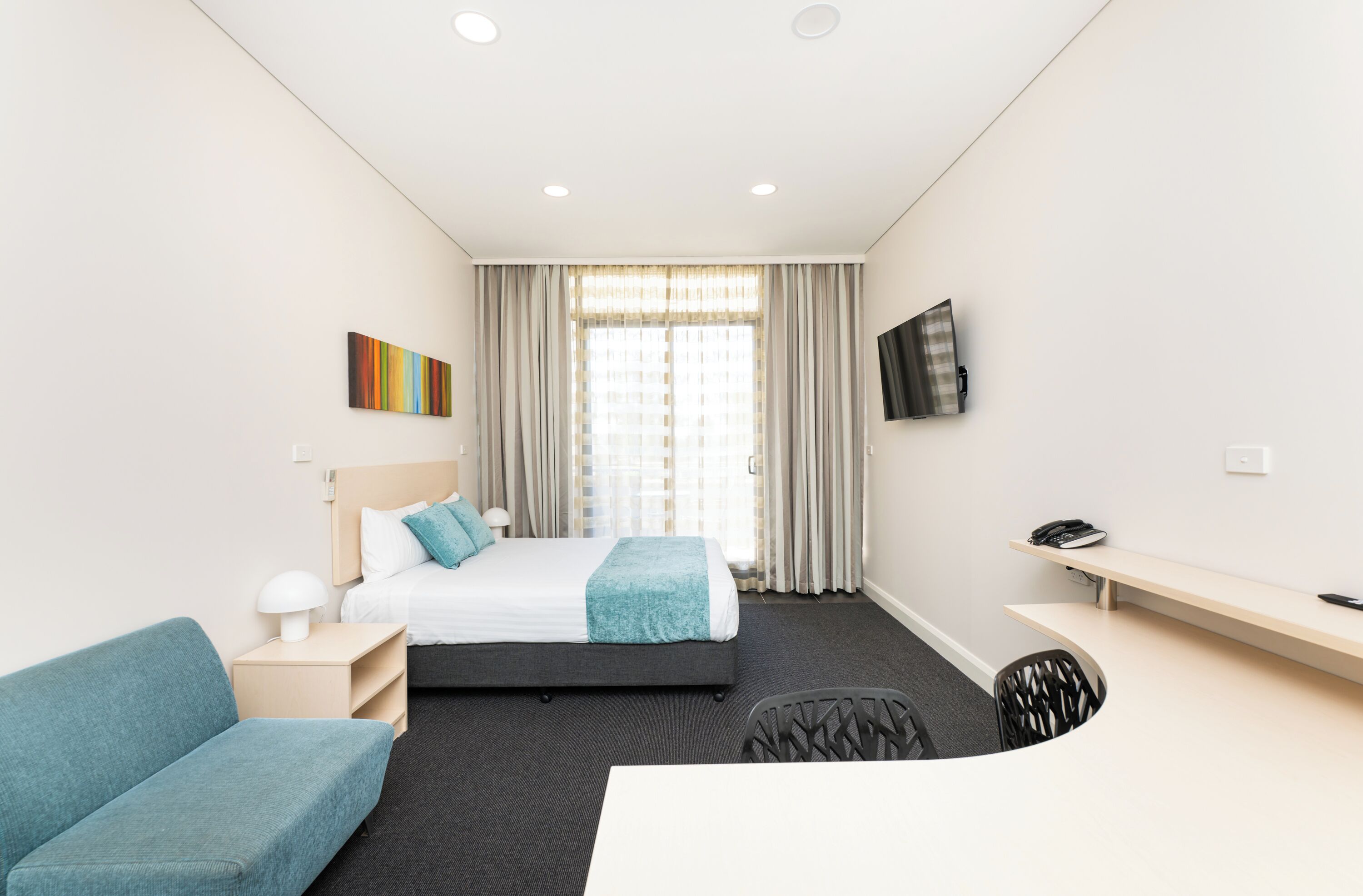 A modern hotel room with a bed, a desk, a chair, a TV, and a painting on the wall.
