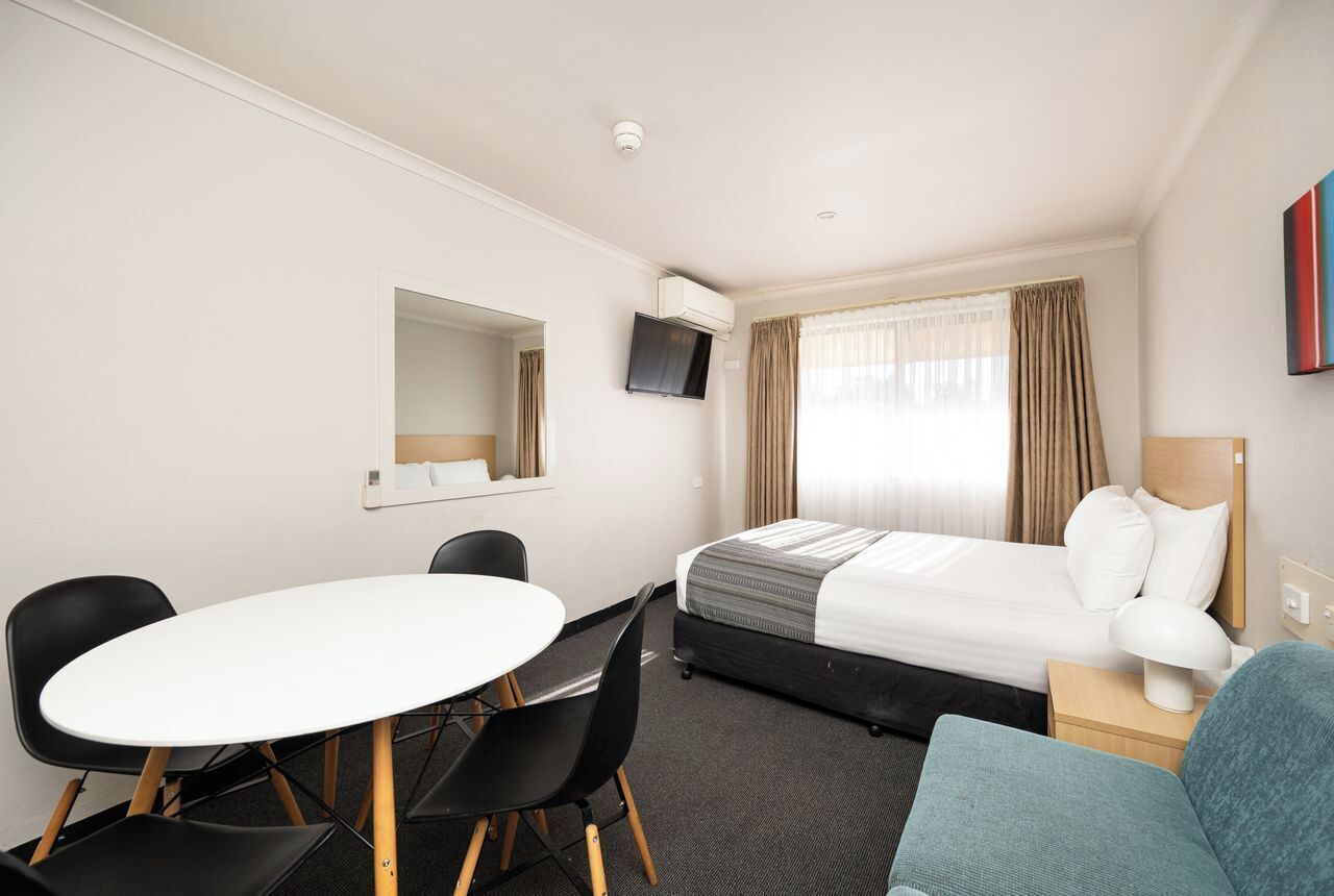 A hotel room with a bed, a desk with chairs, a TV, and a window with curtains.