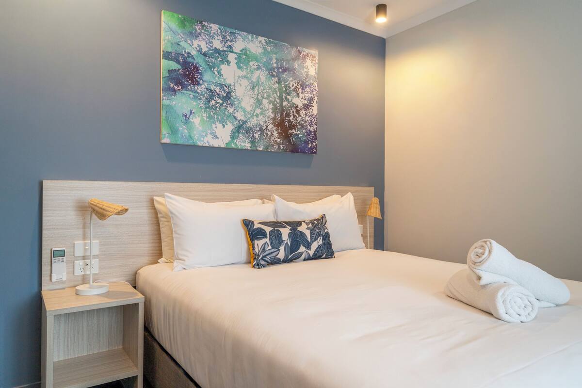 A neatly made bed with white linens, a blue floral pillow, and a rolled towel on the side.