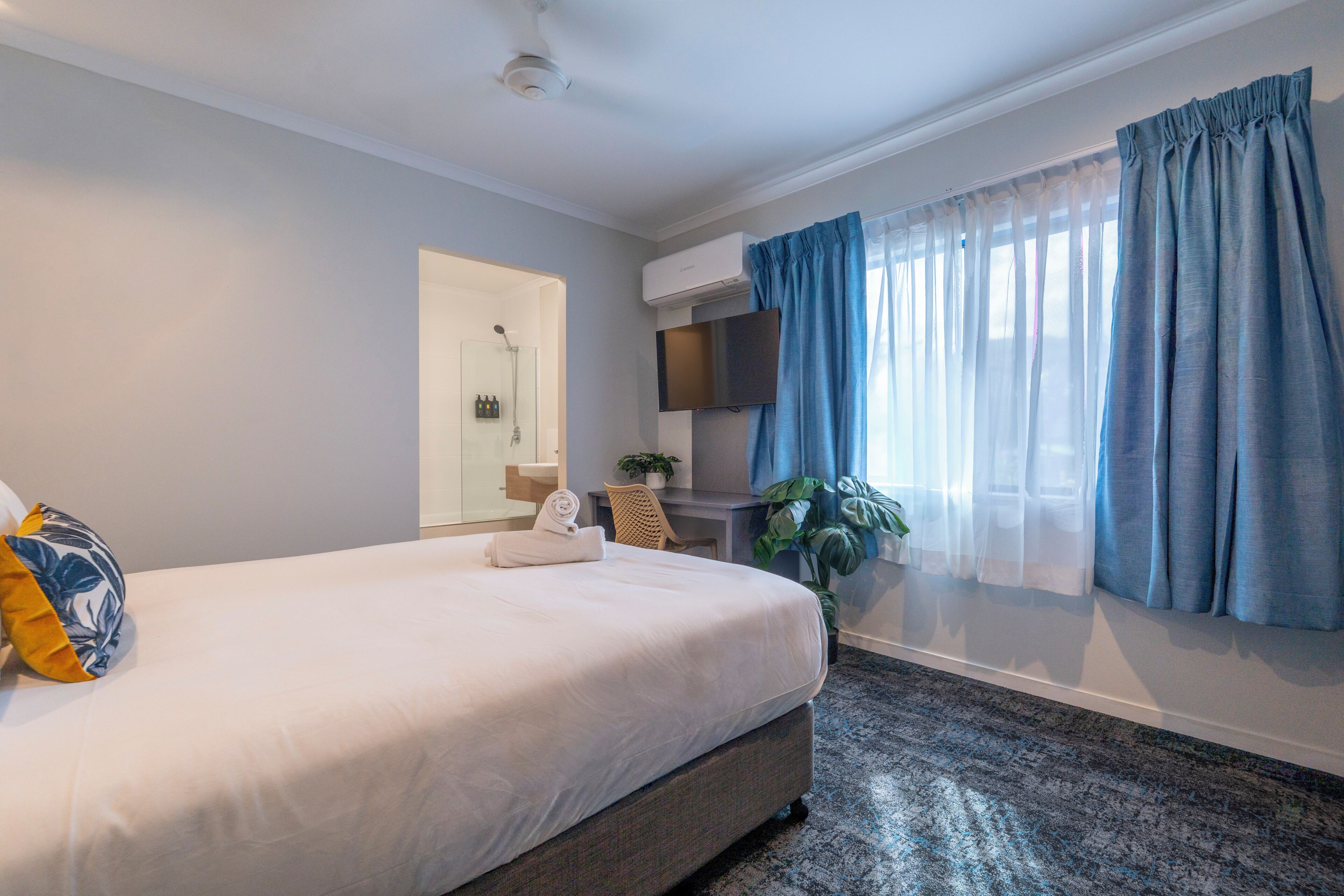 A hotel room with a bed, a desk with a chair, a TV, a shower, and a window with curtains.