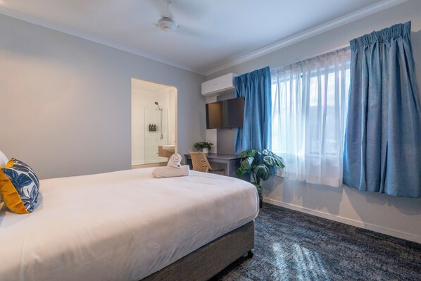 A hotel room with a bed, a desk with a chair, a TV, a shower, and a window with curtains.