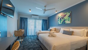 A hotel room with a bed, a desk, a chair, a ceiling fan, and a painting on the wall.
