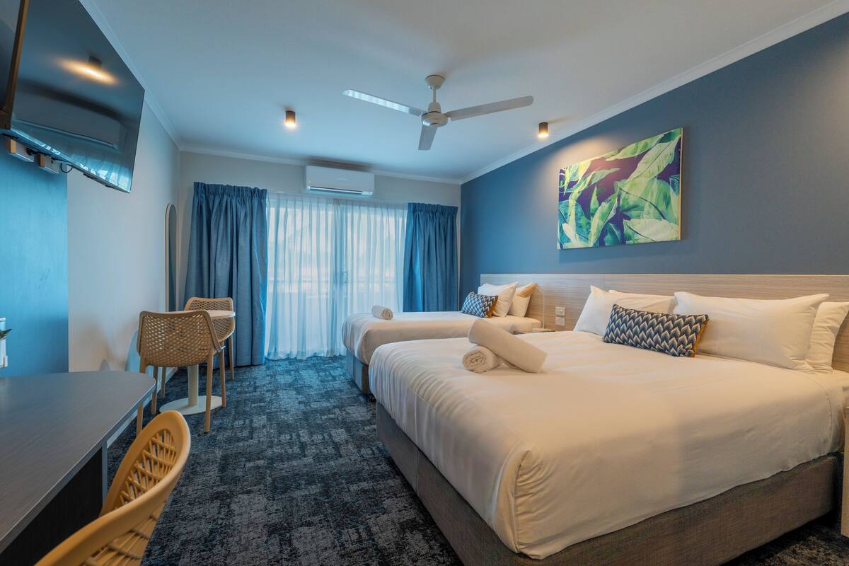 A hotel room with a bed, a desk, a chair, a ceiling fan, and a painting on the wall.