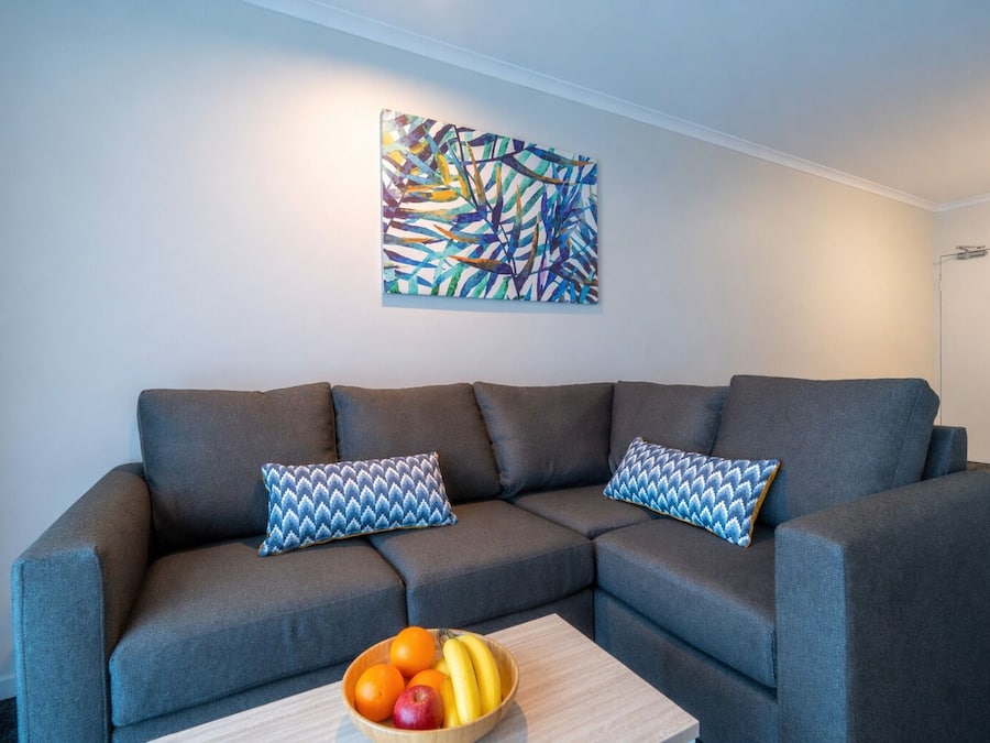 A gray sectional sofa with patterned pillows, a wooden coffee table with fruit, and a colorful abstract painting on the wall.