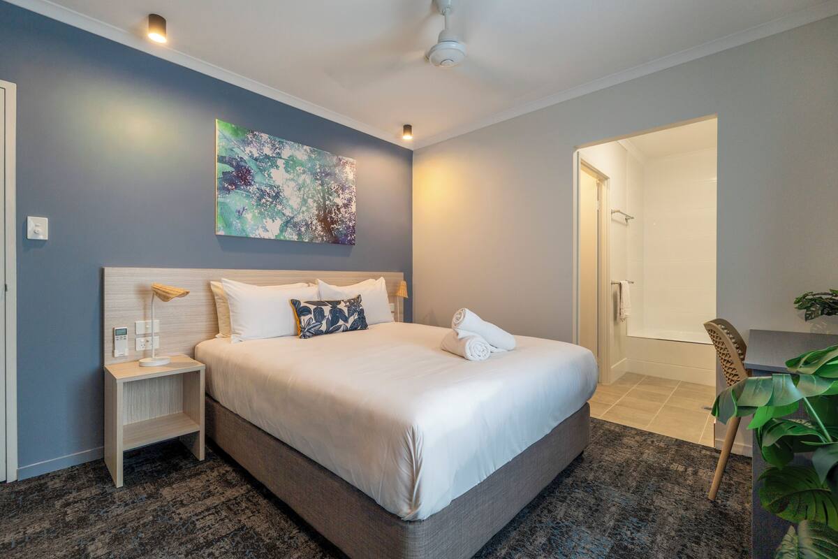 A modern hotel room with a large bed, a bedside table with a lamp, a wall-mounted abstract painting, and a door leading to a bathroom.