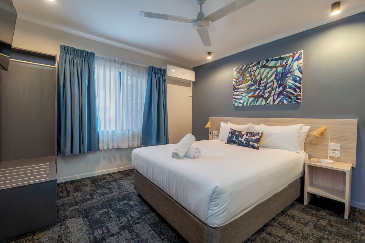 A hotel room with a large bed, a ceiling fan, an air conditioning unit, a nightstand with a lamp, and a window with curtains.